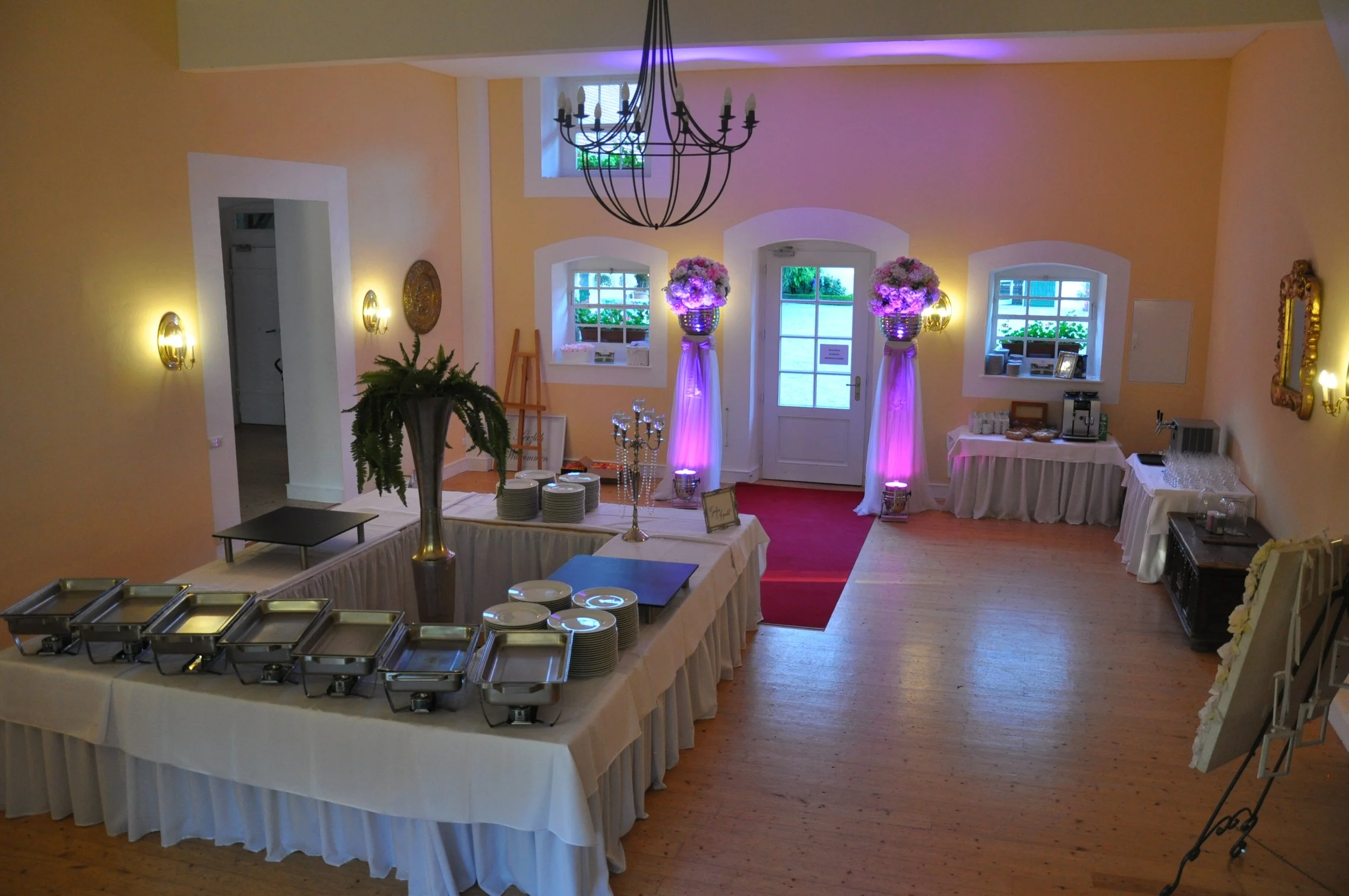 Ein festlich dekorierter Raum mit weißen Bistrotischen, Blumenarrangements auf hohen Ständern, einem Wandspiegel, warm gelben Wänden und einem weißen Buffet mit Geschirr, sowie einer Beleuchtung mit violettem Licht.
