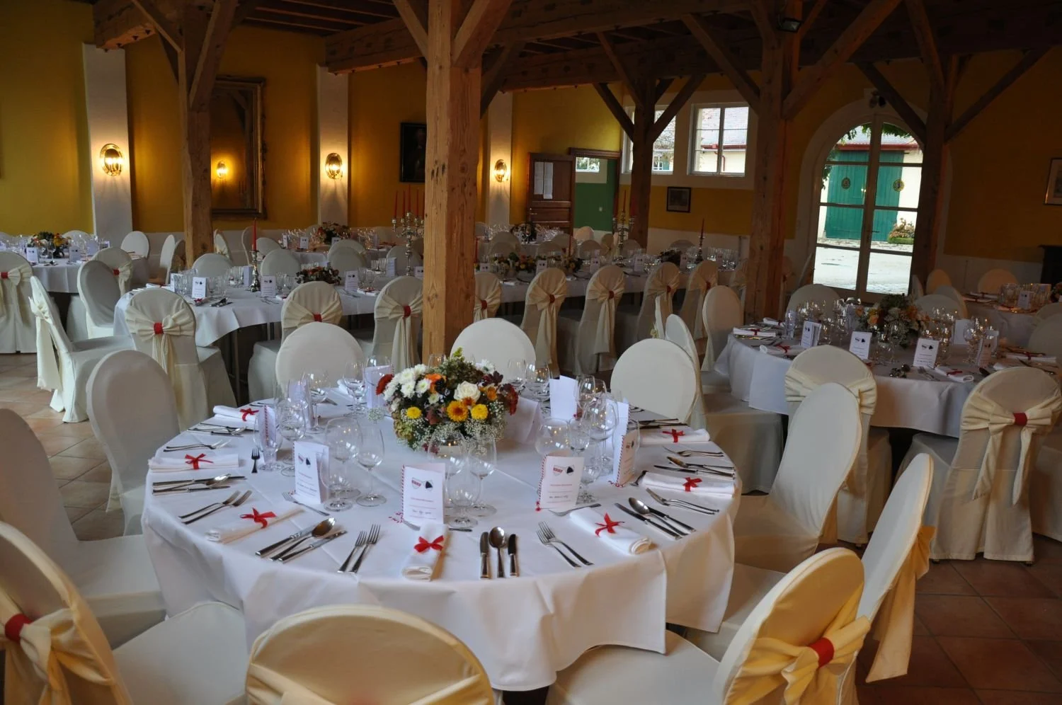 Ein festlich gedeckter Raum mit runden Tischen, weißen Stuhlhussen mit roten Schleifen, Blumenarrangements und eleganter Tischdekoration, bereit für eine Veranstaltung.