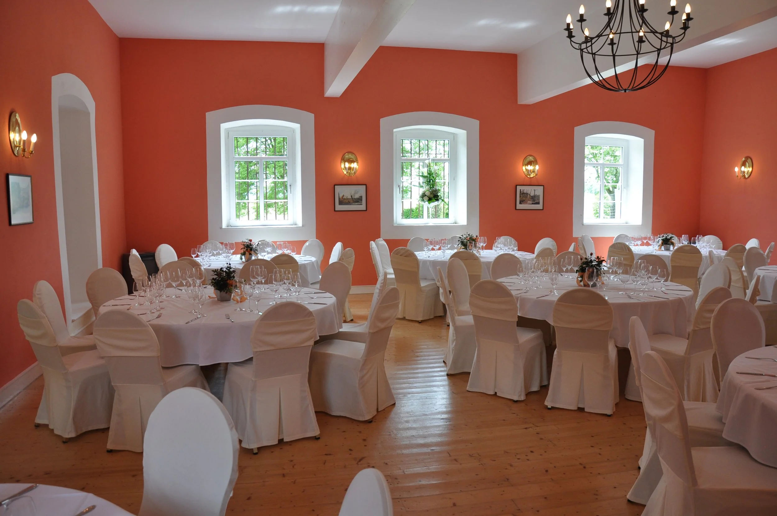 Ein festlich gedeckter Speisesaal mit weißen Tischen und Stühlen, verziert mit kleinen Blumenarrangements. Der Raum hat orange Wände, große Fenster mit Blick auf einen grünen Außenbereich, Wandlampen und eine große elegante Deckenleuchte.