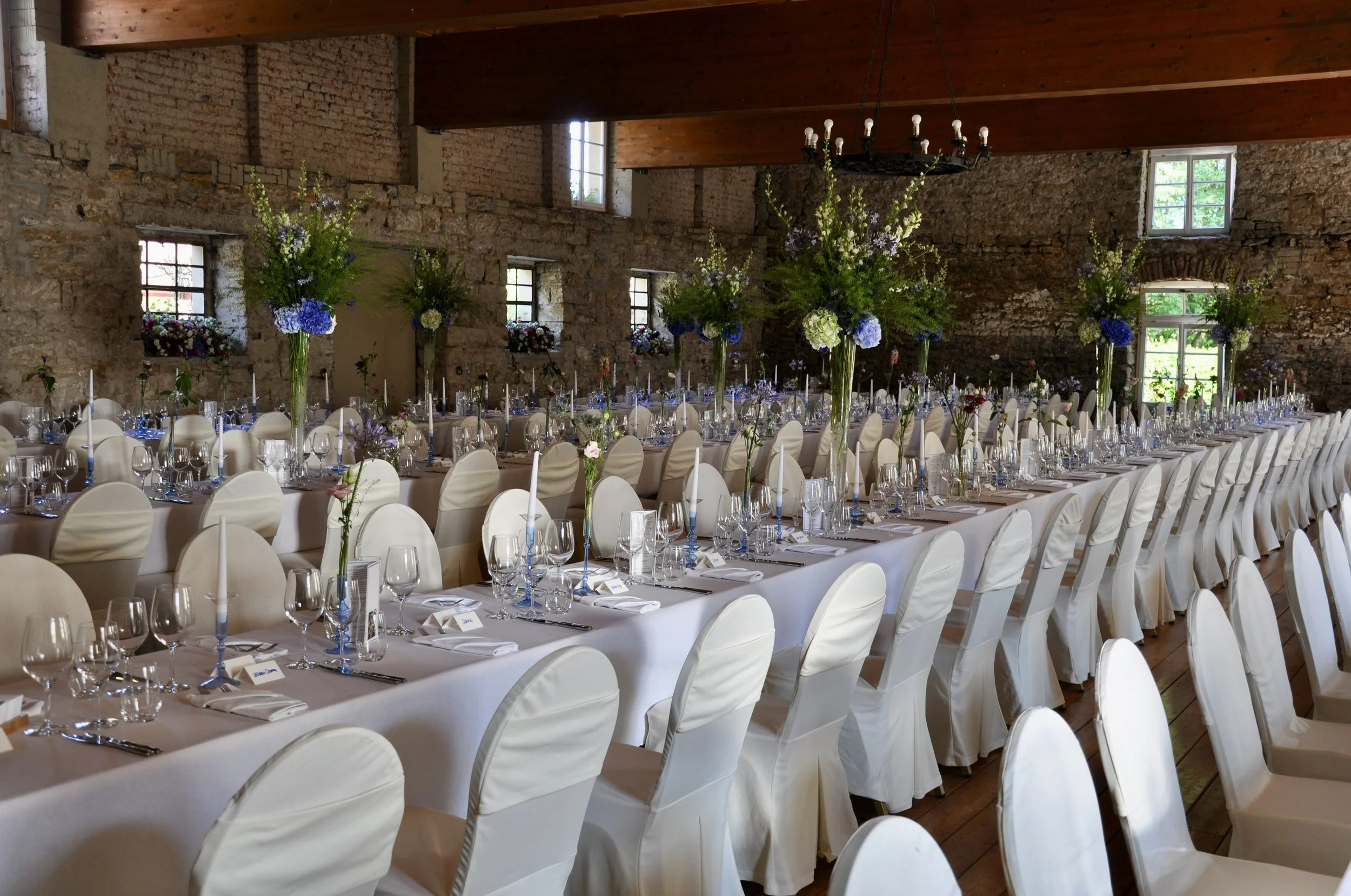 Ein festlich gedeckter Saal mit langen Tischen, weißen Stuhlhussen, Kerzen und großen Blumenarrangements, in einem rustikalen Raum mit Backsteinwänden, Holzbalkendecke und mehreren Fenstern.