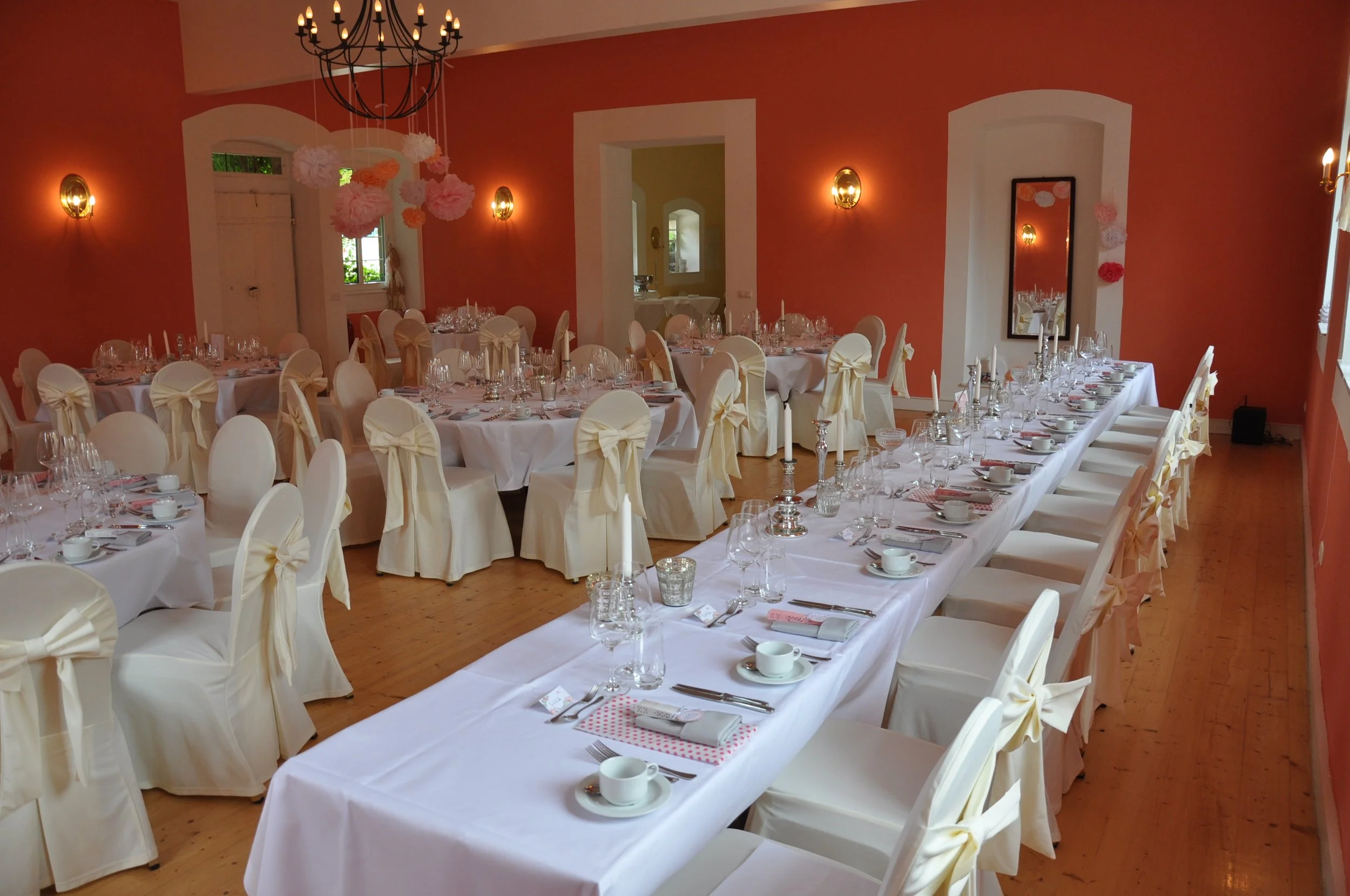 Elegant gedeckter Saal für eine Feier mit runden Tischen, weißen Stuhhussen mit Schleifen, Kerzen, Gläser und Tassen, beige und rosa Dekorationen, Chandelier und goldene Wandlampen, warme Beleuchtung, Holzboden, in einem festlichen Raum