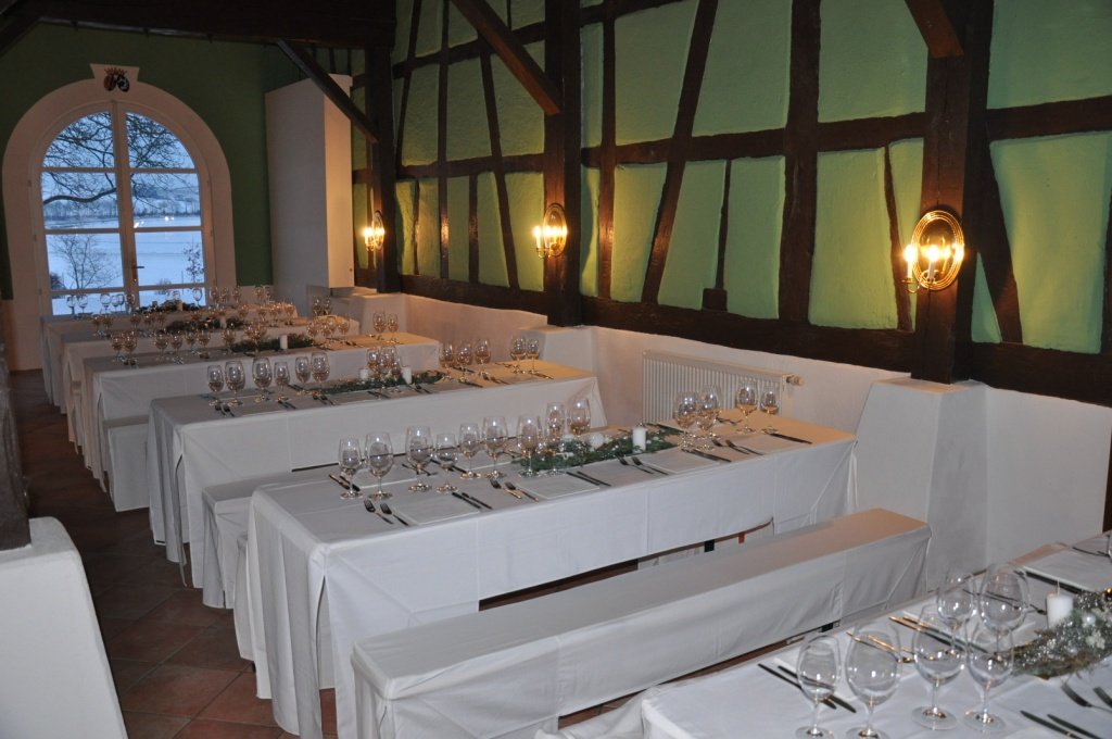 Festlich gedeckte Tische in einem Weihnachtsessen-Restaurant mit weißen Tischtüchern, Kristallgläsern und silbernem Besteck, Weihnachtsdeko in der Mitte, große Fenster mit Blick auf schneebedeckten Baum.
