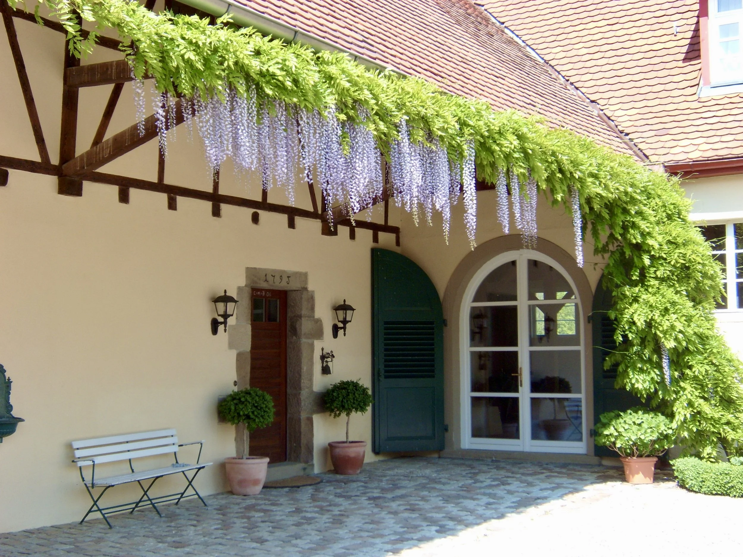 Ein Haus mit einer Veranda, grünen Türen, Vasen mit Pflanzen, einer Bank und rankendem Flieder an der Fassade.