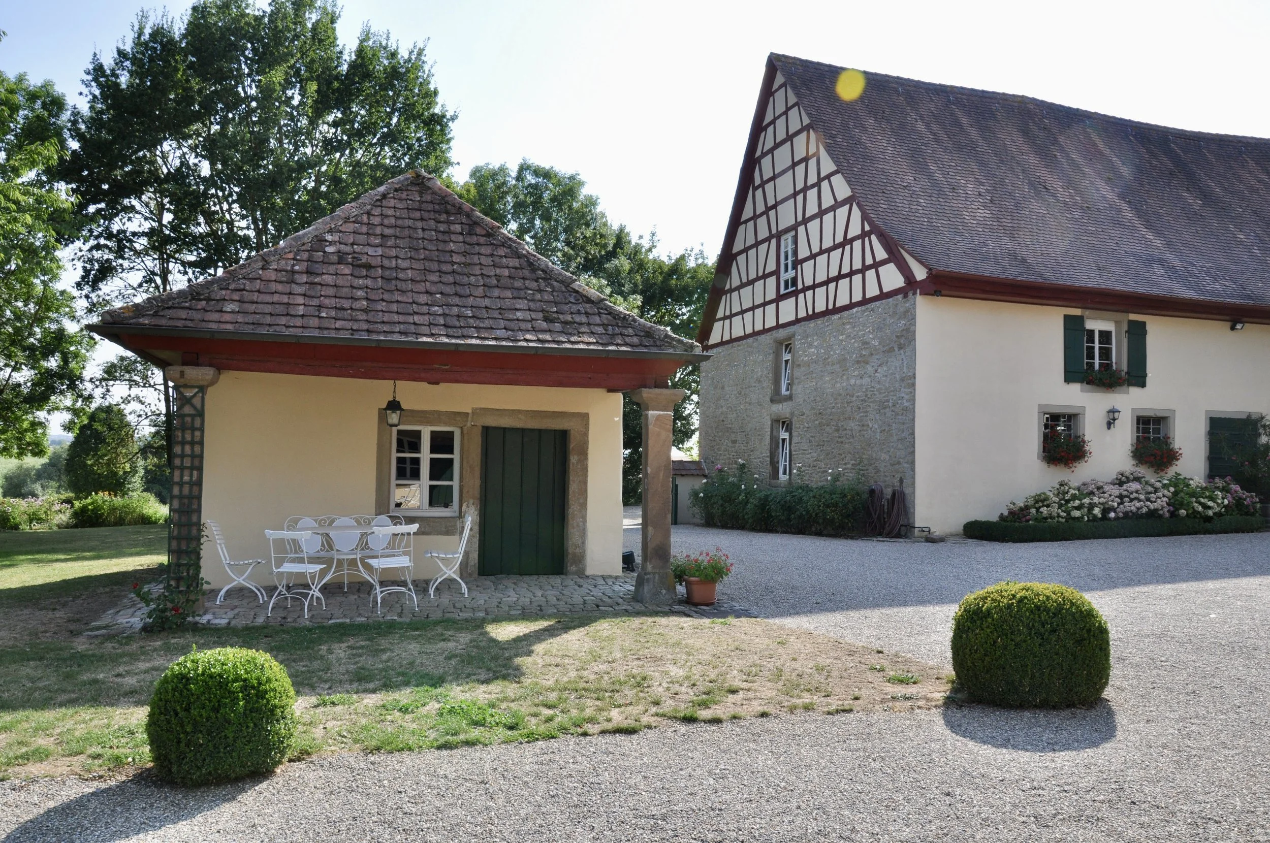 Ein altes Gebäude mit einem steinernen Untergeschoss und einer Fachwerkwand, umgeben von einem Garten mit niedrigen, runden Hecken, einem großen Baum und einer Terrasse mit weißen Stühlen und einem Tisch.
