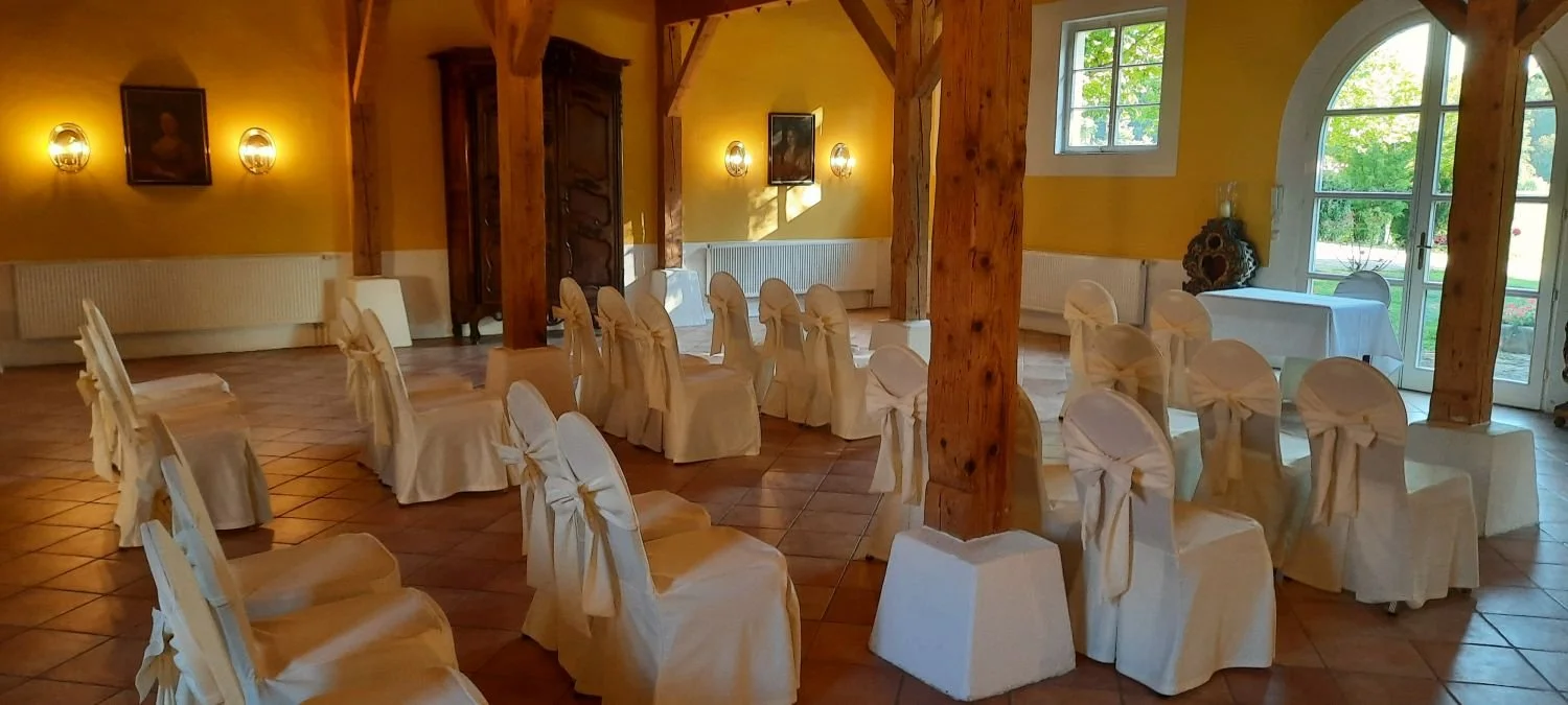 Ein festlich dekorierter Saal mit weißen Stuhlhussen, die mit Schleifen gebunden sind. Der Raum hat große Fenster und Holzträger, mit Wanddekoration und warmem Licht.