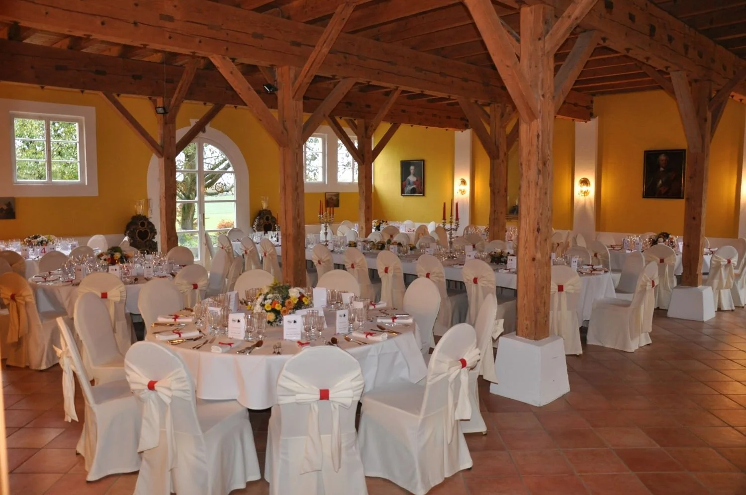 Der festlich gedeckte Gartensaal mit runden Tischen, weißen Stuhlhussen, Blumenarrangements, Kerzen und Besteck, dekoriert für eine Feier, mit Holzbalken im Deckenbereich und gelben Wänden.