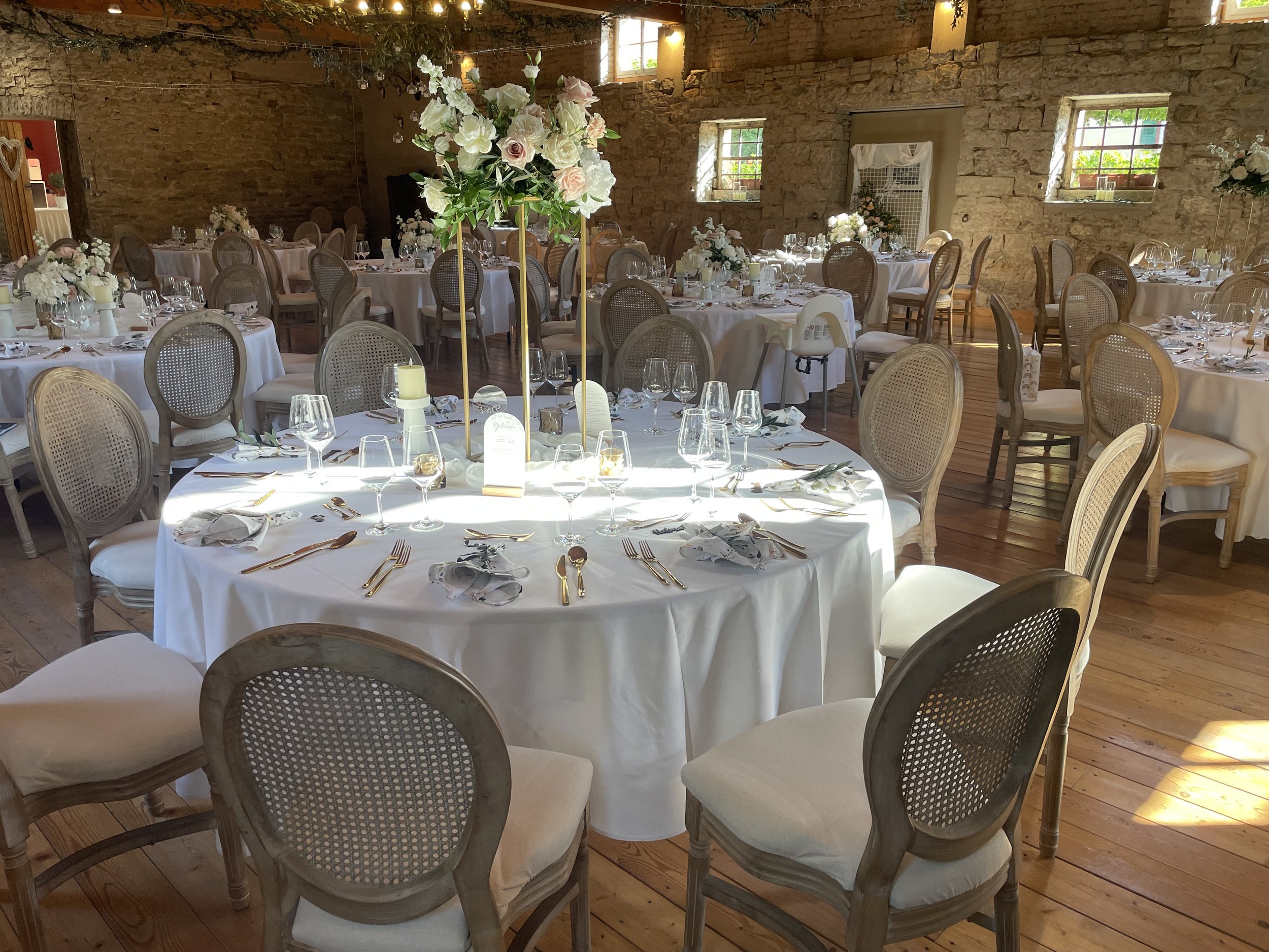 Ein festlich gedeckter Saal mit mehreren runden Tischen, weißen Tischdecken, Weingläsern, Besteck und Papierservietten, zentrale Blumenarrangements mit weißen und rosa Blumen, viele Stühle um die Tische herum, rustikale Steinwände, kleine Fenster, na