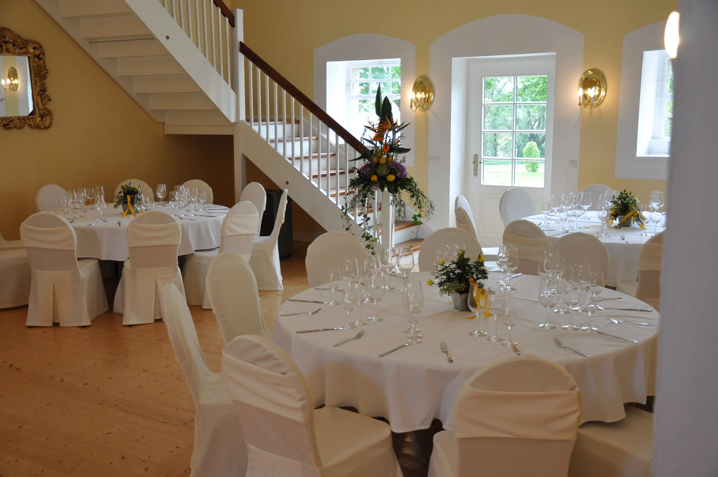 Der elegante Gelbe Saal auf Gut Schwarzerdhof mit runden Tischen, weißen Stuhllehnen, geschmackvollen Blumenarrangements und großen Fenstern mit Blick ins Grüne.