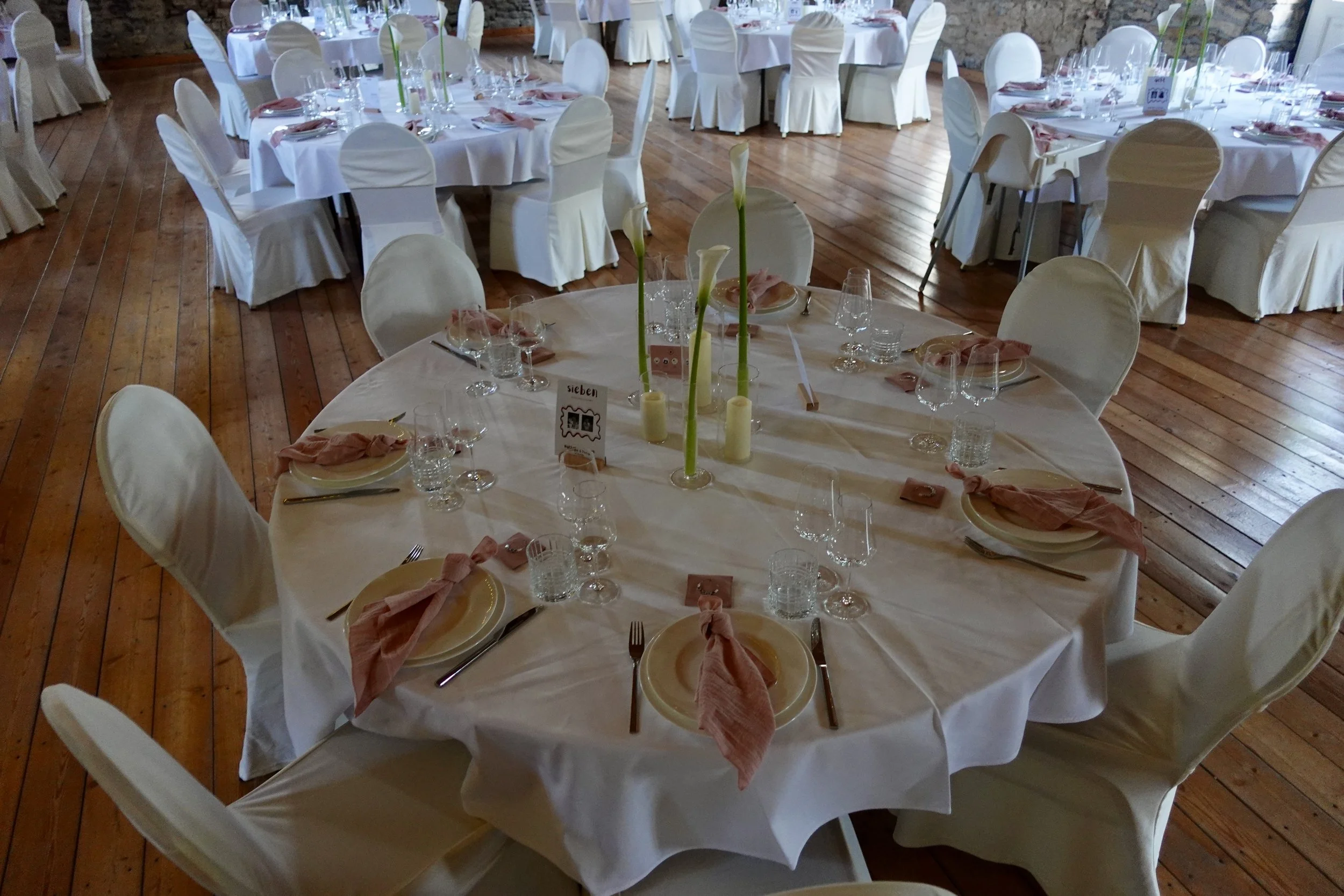 Ein festlich gedeckter Raum mit runden Tischen, die weiß eingedeckt sind, Kerzen und Blumen in hohem Glasvasen als Dekoration. Die Tische sind mit Gläsern, Tellern, Besteck und gefalteten Servietten vorbereitet, die Stühle sind mit weißen Bezügen bed
