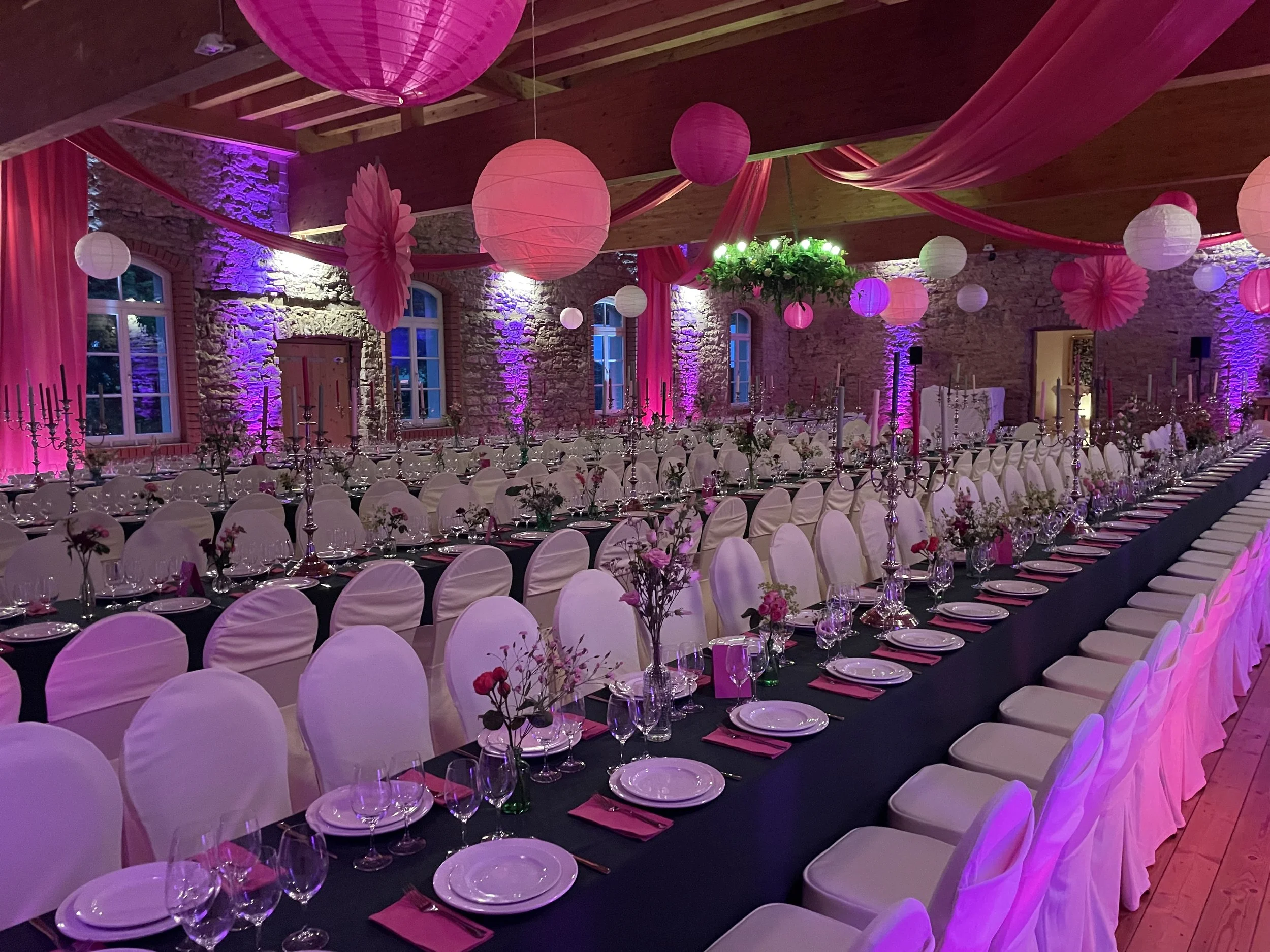 Ein festlich gedeckter Raum mit runden Tischen, weißen Stühlen, Kerzenhaltern, Blumen und rosa Dekorationen, beleuchtet mit bunten Lichtern und Lampions.