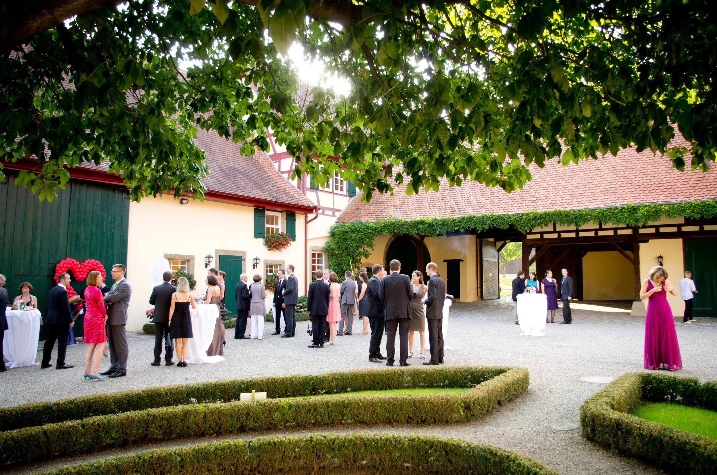 Eine Gruppe von Menschen in formeller Kleidung bei einer Feier auf einem Bauernhof mit historischen Fachwerkhäusern, die im Hintergrund zu sehen sind. Es scheint ein festlicher Anlass zu sein.
