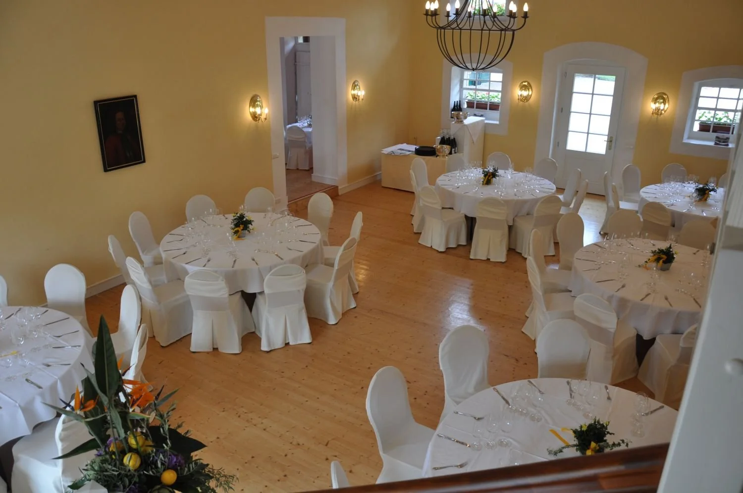 Ein festlich gedeckter Saal mit mehreren weiß eingedeckten Tischen, goldenen Kerzenleuchtern, weißen Stuhlhussen, dekorativen Pflanzen und einem Kronleuchter an der Decke, eingerichtet für eine Feier oder Veranstaltung.