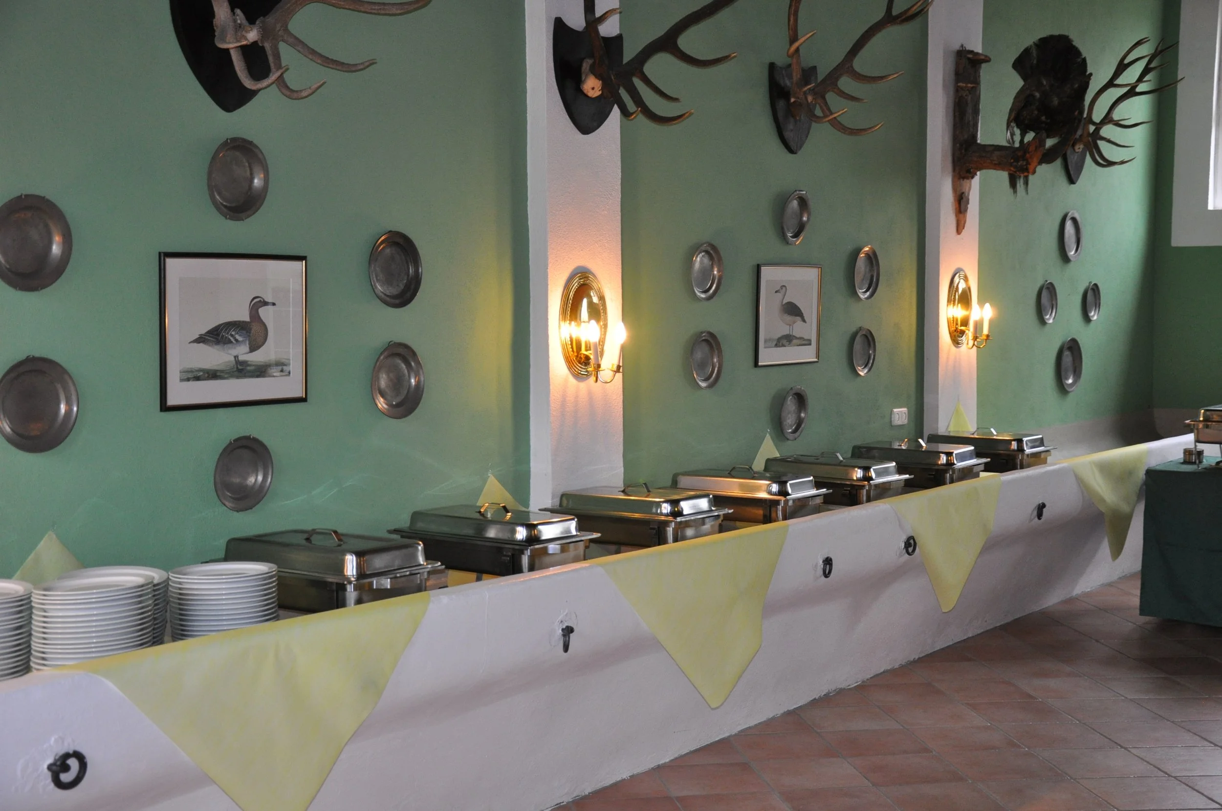 Ein Buffetbereich in einem Restaurant mit grüner Wanddekoration, einschließlich Bilder von Wasservögeln, Wandlampen, dekorativen Tierköpfen und Metallplatten an der Wand. Es gibt mehrere Chafing Dishes auf dem Buffet, sowie einen Stapel weißer Teller