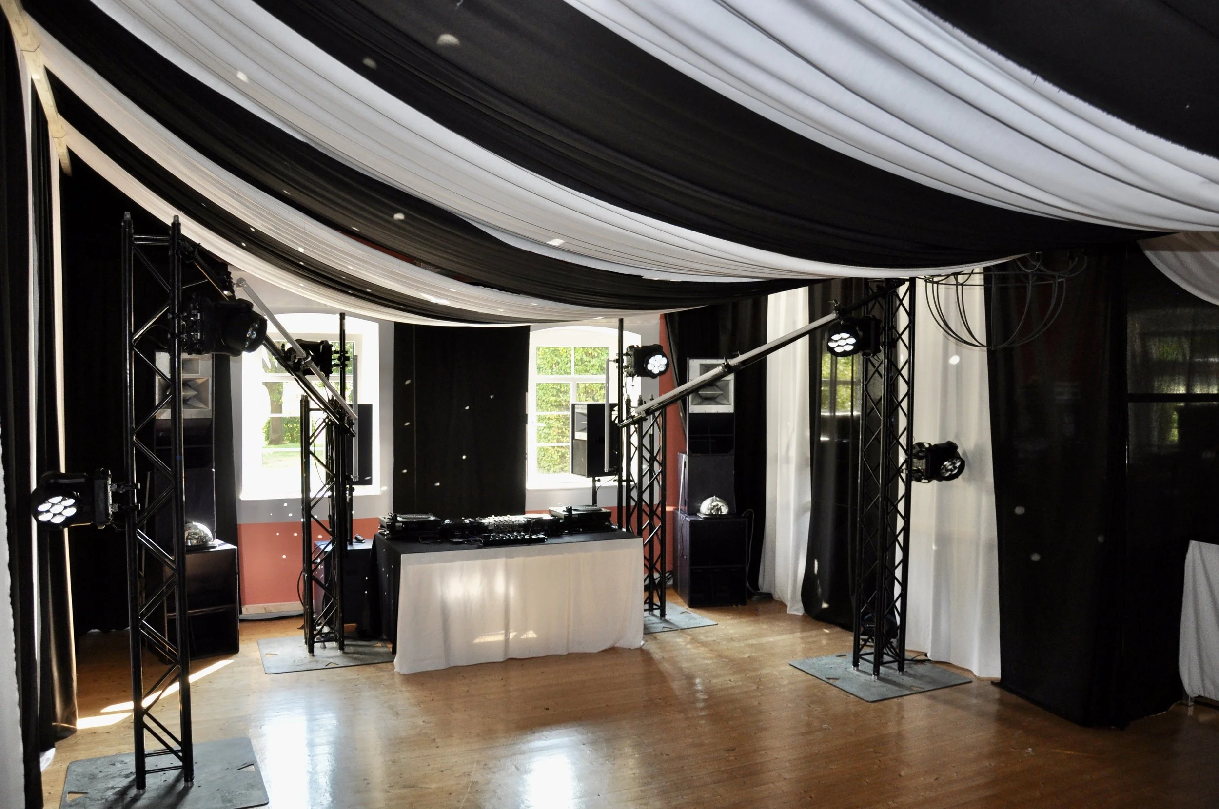 DJ-Setup mit Lautsprechern, Beleuchtung und Mikrofon auf einer Eventbühne in einem festlich dekorierten Raum mit Vorhängen und großen Fenstern.