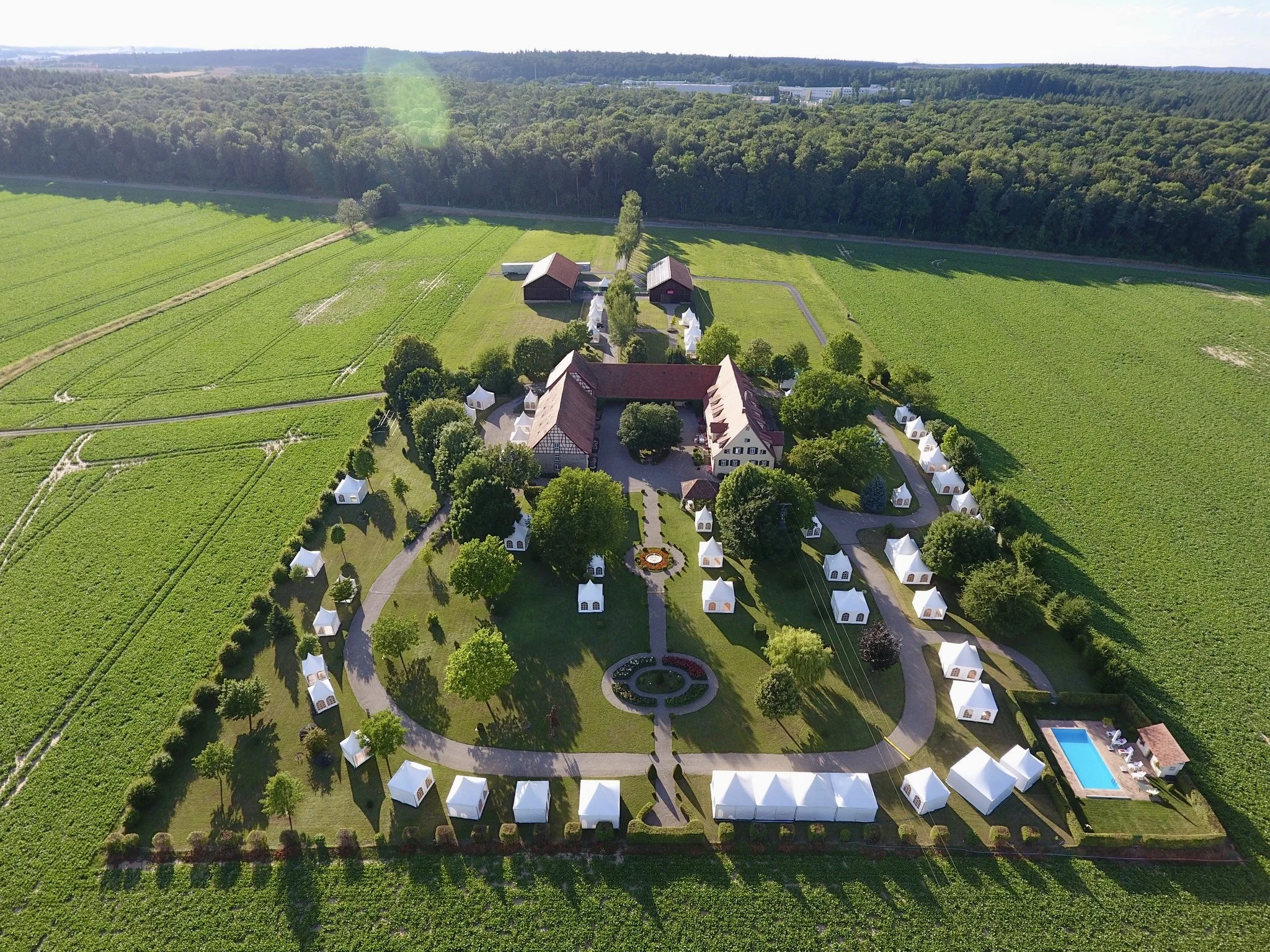 Luftaufnahme eines großen Anwesens mit mehreren Gebäuden, Garten und umgebenden Feldern, umgeben von Wäldern