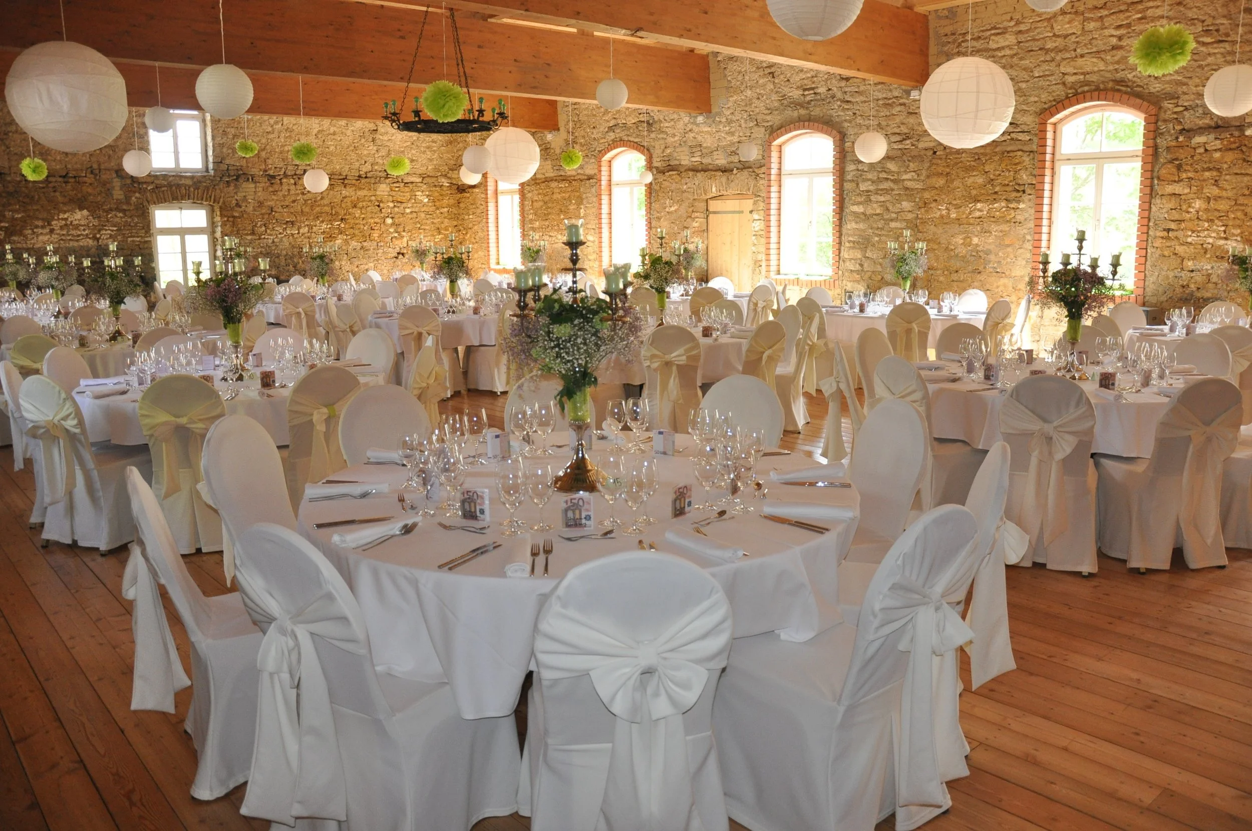 Ein festlich gedeckter Saal mit runden Tischen, weißen Tischdecken, Stuhlhussen mit Schleifen, mehreren Weingläsern, Kerzenleuchtern und Blumensträußen, in einem Raum mit Steinwänden, großen Fenstern und hölzernen Deckenbalken, dekoriert für eine Fei