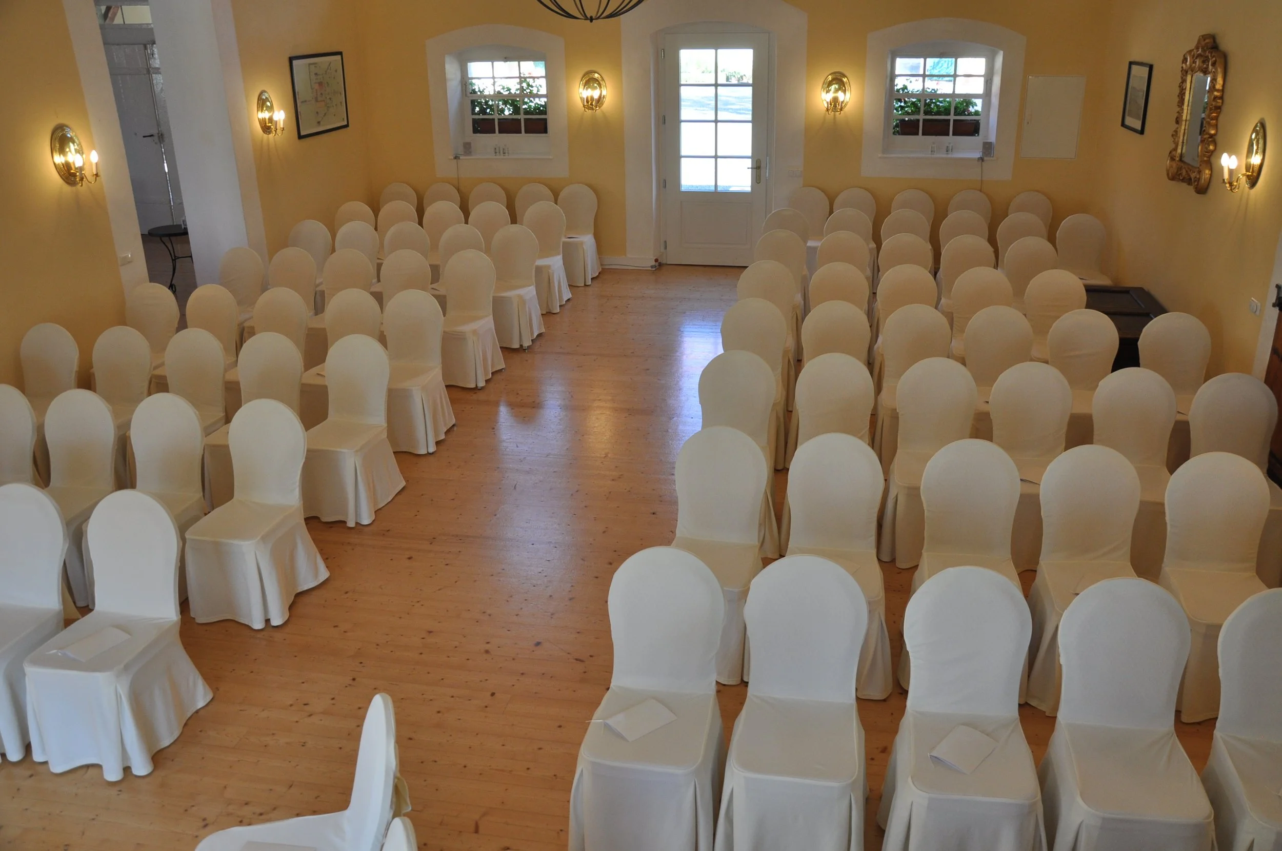 Ein Saal mit vielen weißen, bezogenen Stühlen, die für eine Veranstaltung vorbereitet sind. Der Raum hat gelbe Wände, Holzboden, zwei Fenster und eine Tür im Hintergrund. Es gibt Wandlampen und einen großen, goldfarbenen geschwungenen Spiegel an der 