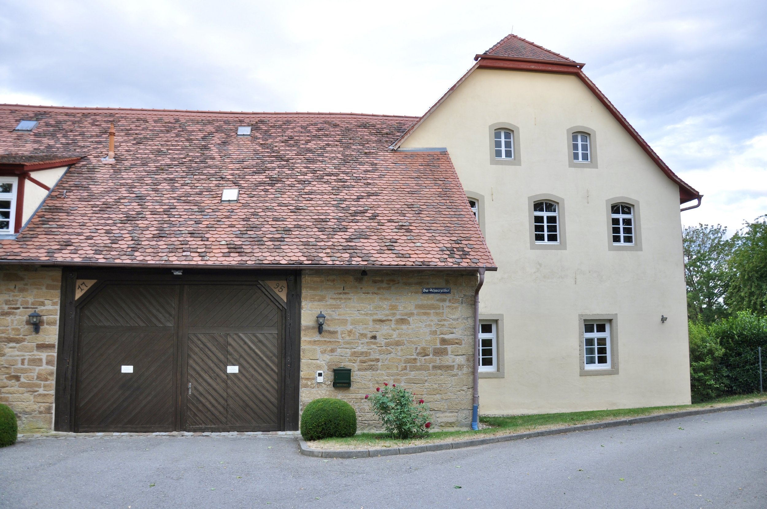 Ein Haus mit Ziegeldach, weißen Fenstern und einem großen Holztor vor einer steinernen Fassade, mit Garten und einem Straßenzugang im Vordergrund.