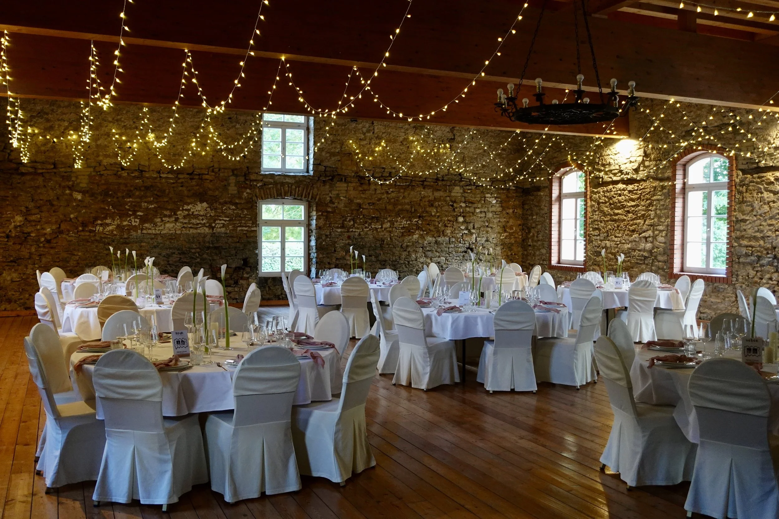 Ein festlich dekorierter Veranstaltungsraum mit runden Tischen, weißen Stuhlhussen, Tellern, Gläsern und Blumenarrangements, festliche Lichter und eine Barockkronleuchte hängen von der Holzdecke. Große Fenster lassen natürliches Licht herein.