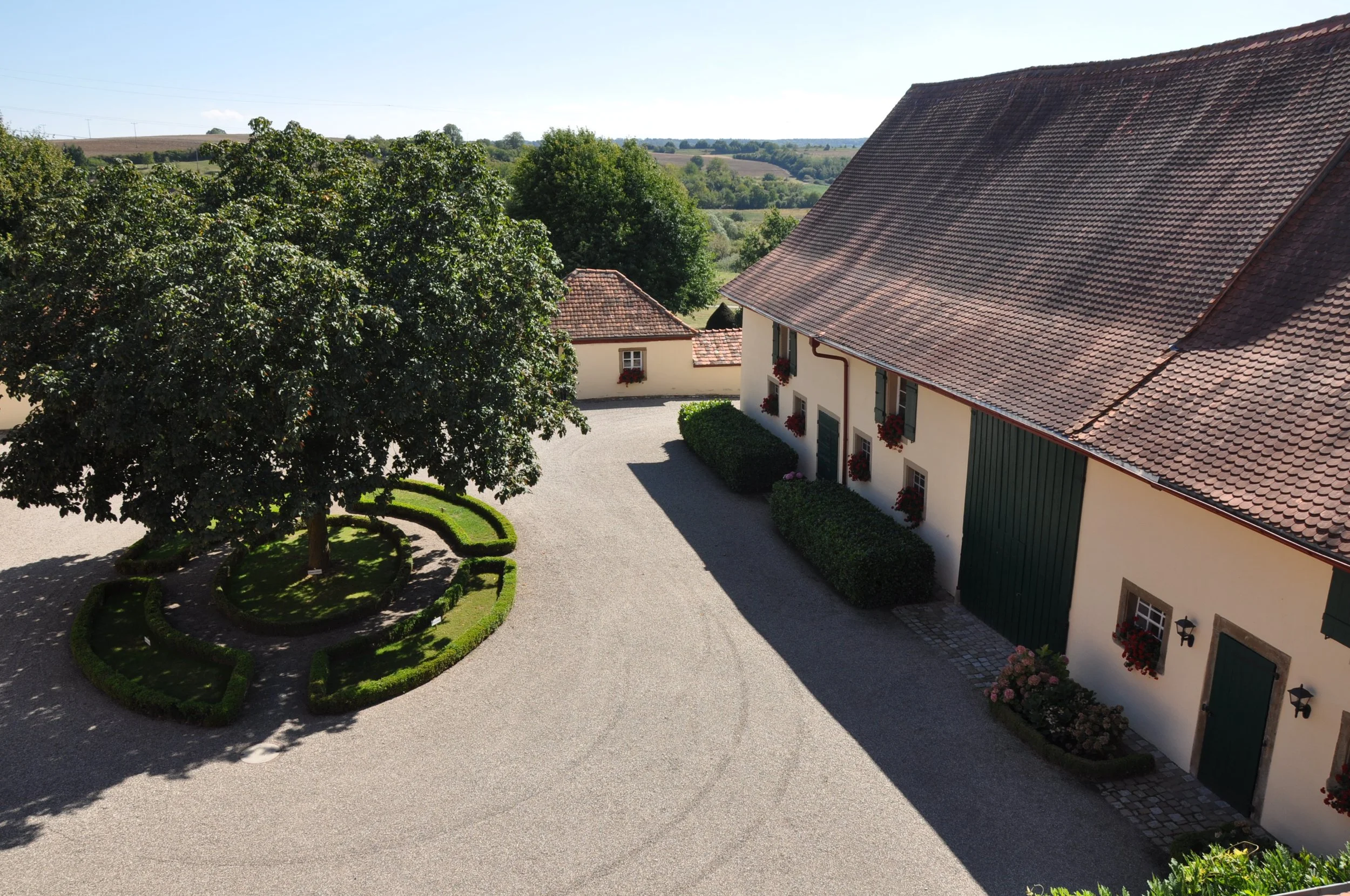 Ein Hof mit zwei Gebäuden, einem gepflegten Garten mit Hecken und einem großen Baum, umgeben von einer ländlichen Landschaft mit Feldern und Hügeln im Hintergrund.