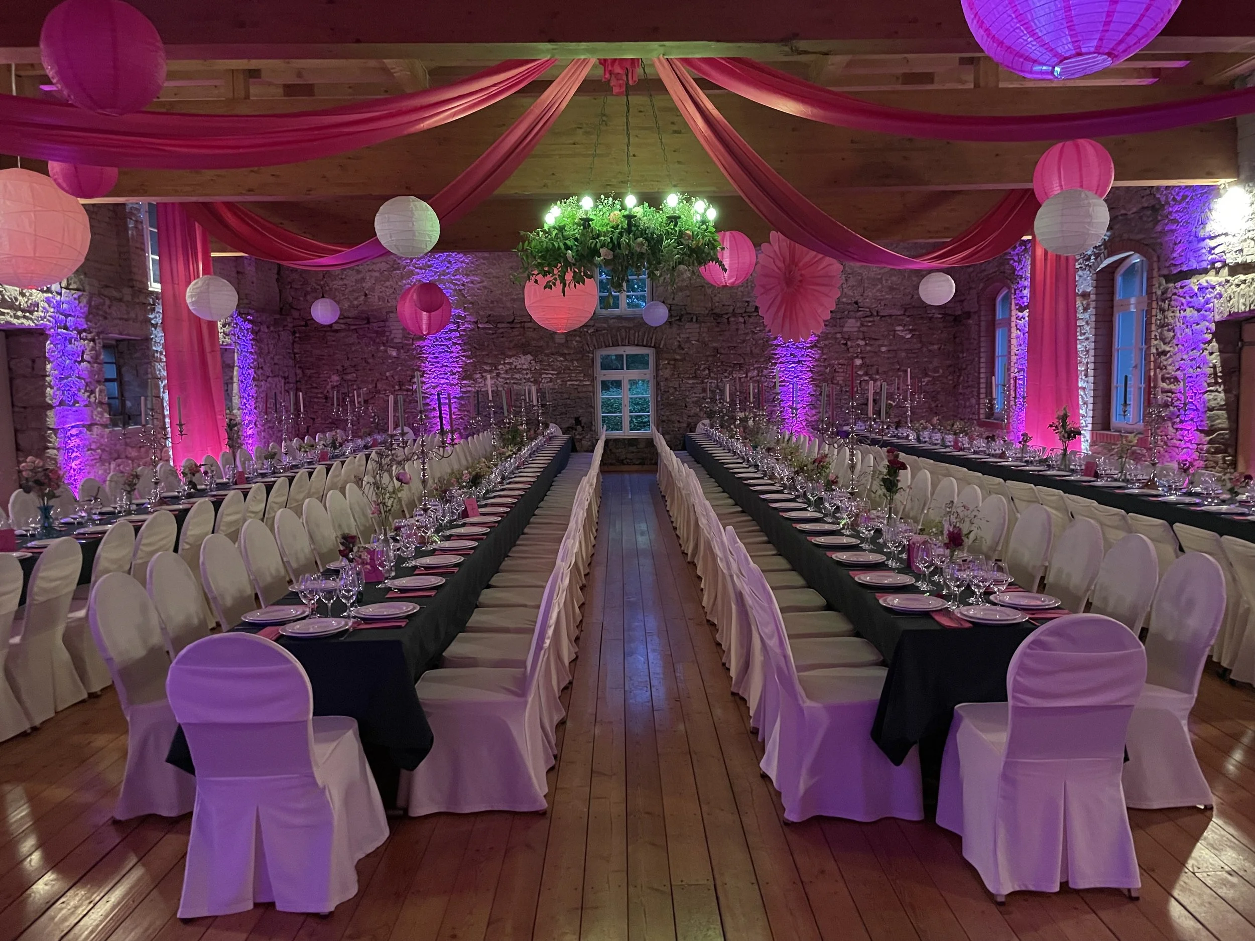 Ein festlich dekorierter Saal mit langen Tischen, weißen Stühlen, pinken und weißen Papierlampions, pinken Stoffen an der Decke und bunten Lichtprojektionen auf den Steinwänden.