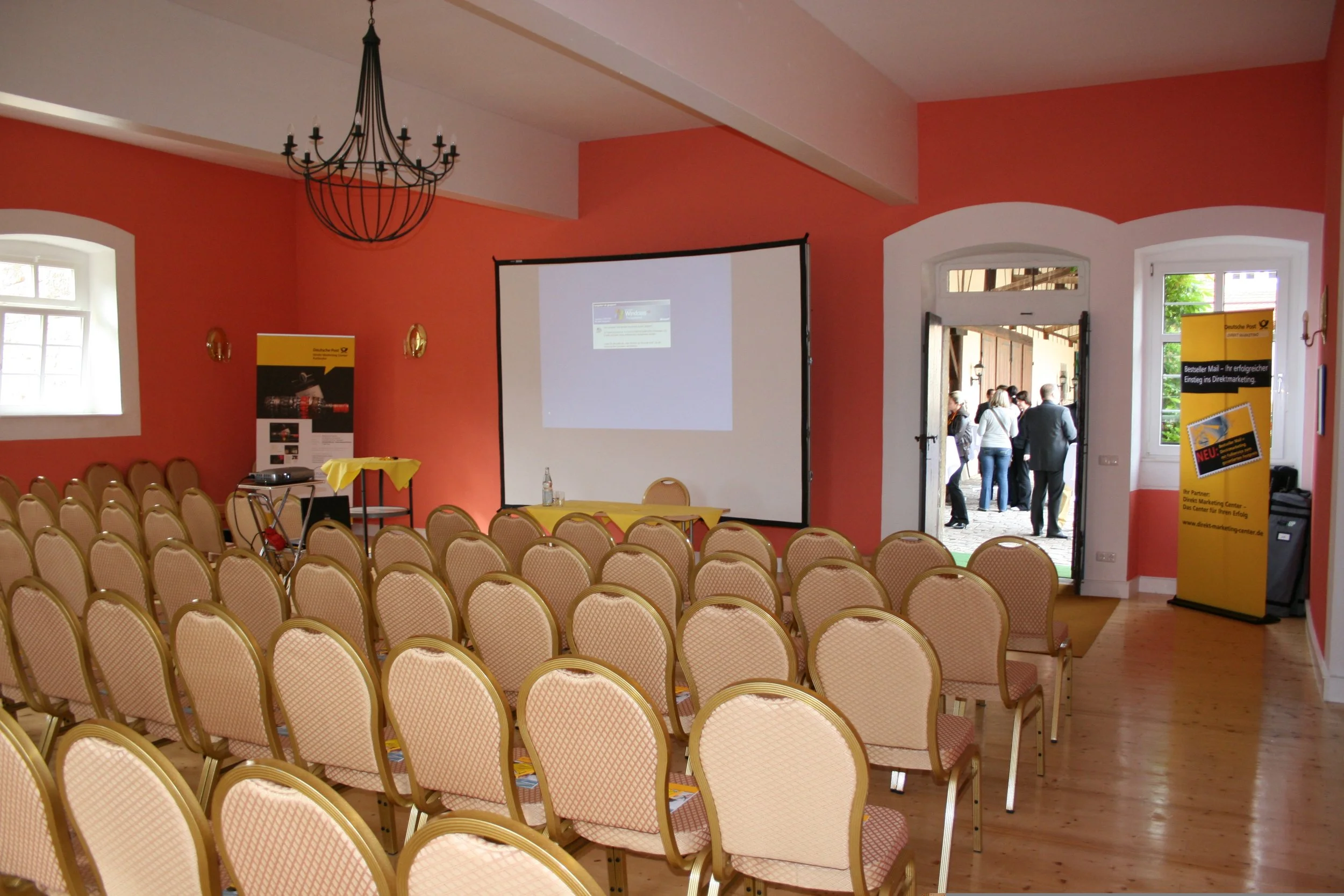Ein Veranstaltungsraum mit vielen leeren Stühlen, einem large weißen Projektorbildschirm, und einem stilvollen Kronleuchter an der Decke. Es gibt auch einige Werbestände an den Wänden.