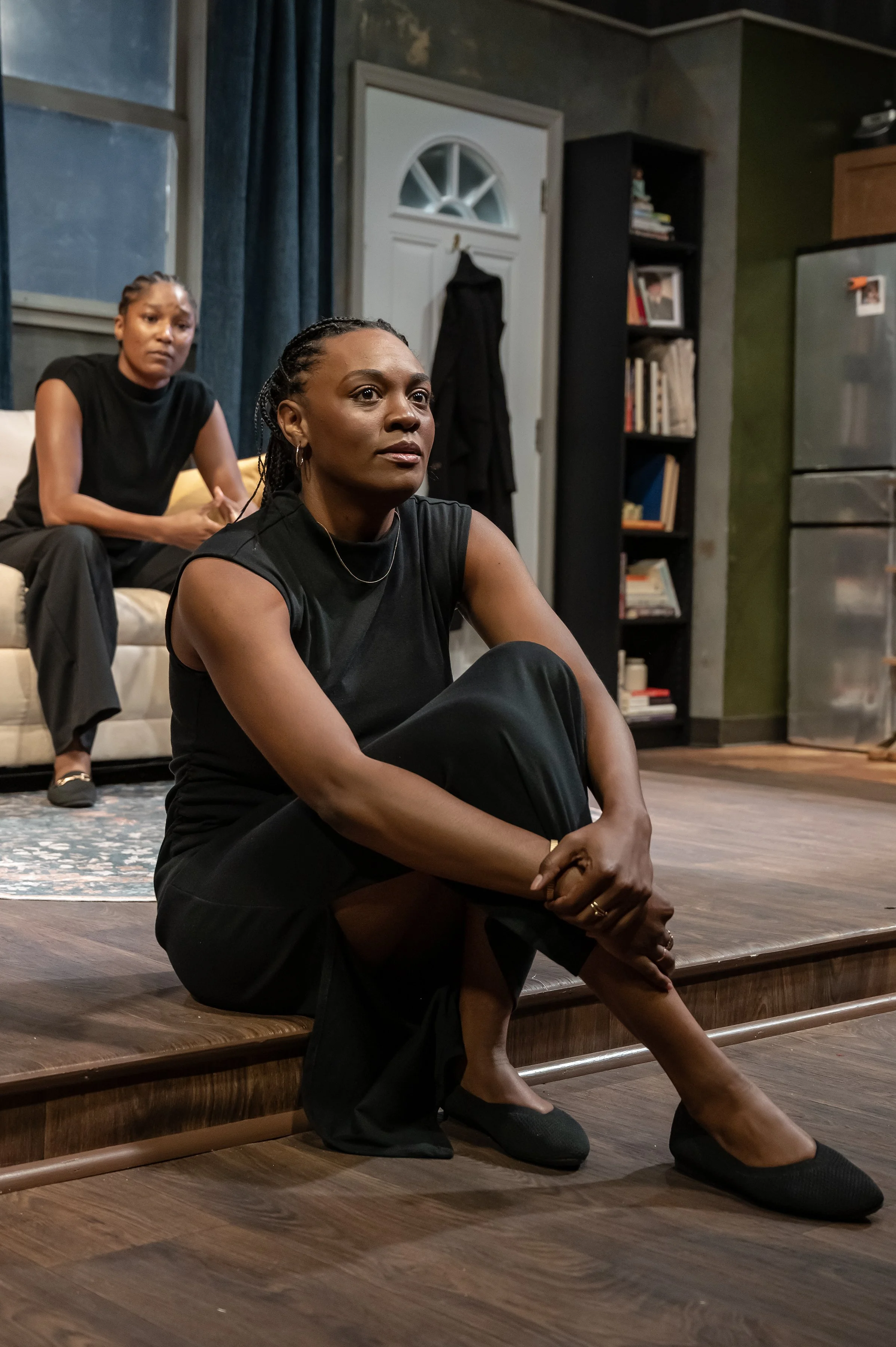 Close-up of Mina and Sade in front of the living room and kitchen dressed sets in the play “Furlough’s Paradise” by A.K. Payne