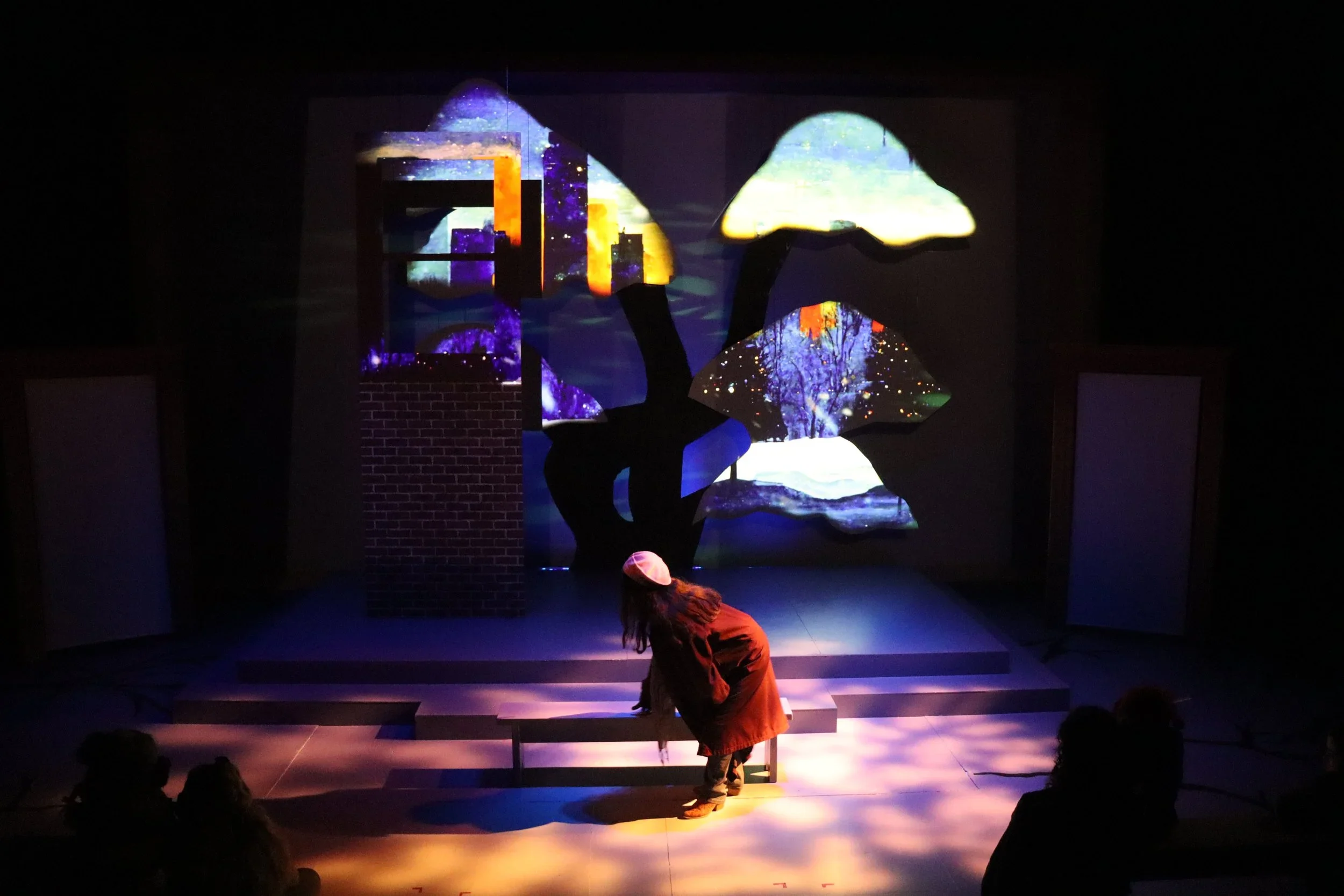 Night scene picturing Rebecca laying down on a bench in front of a tree with projected winter images for the play “The Long Christmas Ride Home” by Paula Vogel