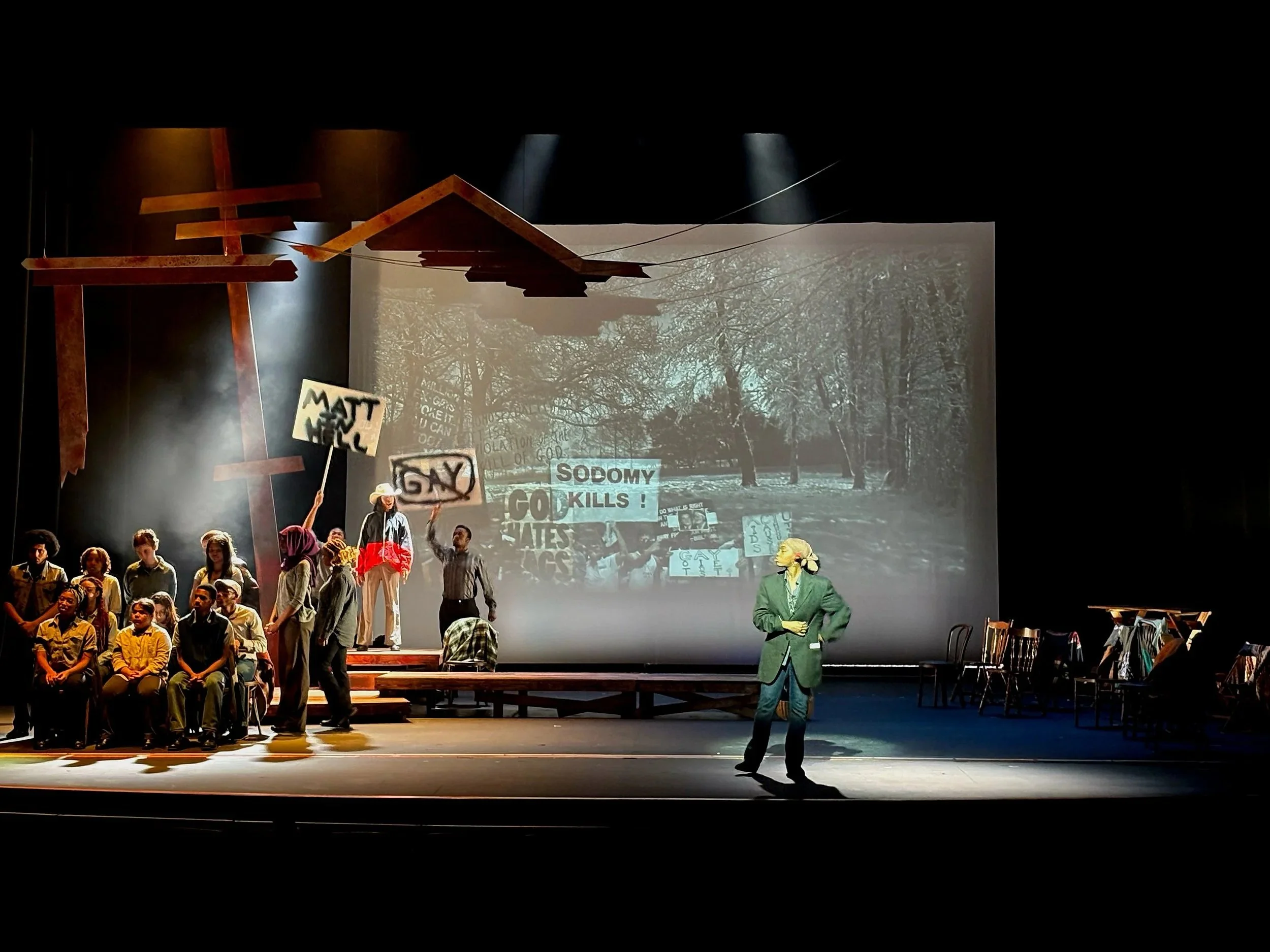 Cast onstage at a anti-LGBTQ rally, emphasized with the projection design for the play “The Laramie Project” by Moises Kaufman and the Tectonic Theater Project