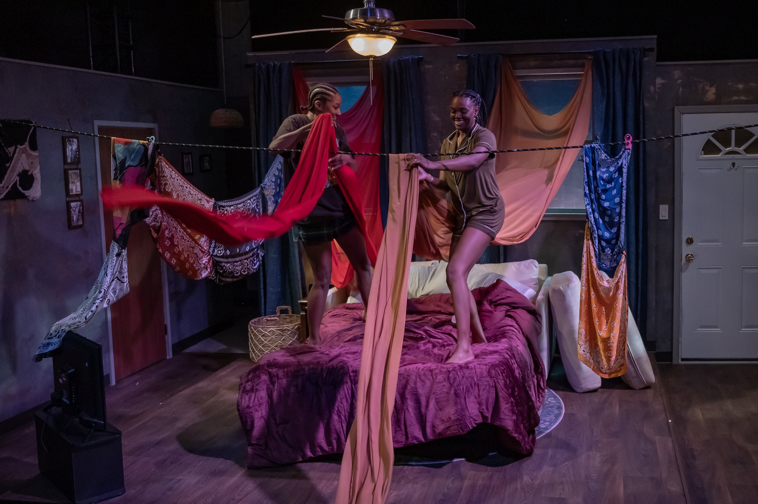 The two main characters standing on a sofa bed and draping fabric from curtains and headscarves over a length of rope to create a fort during the play “Furlough’s Paradise” by A.K. Payne