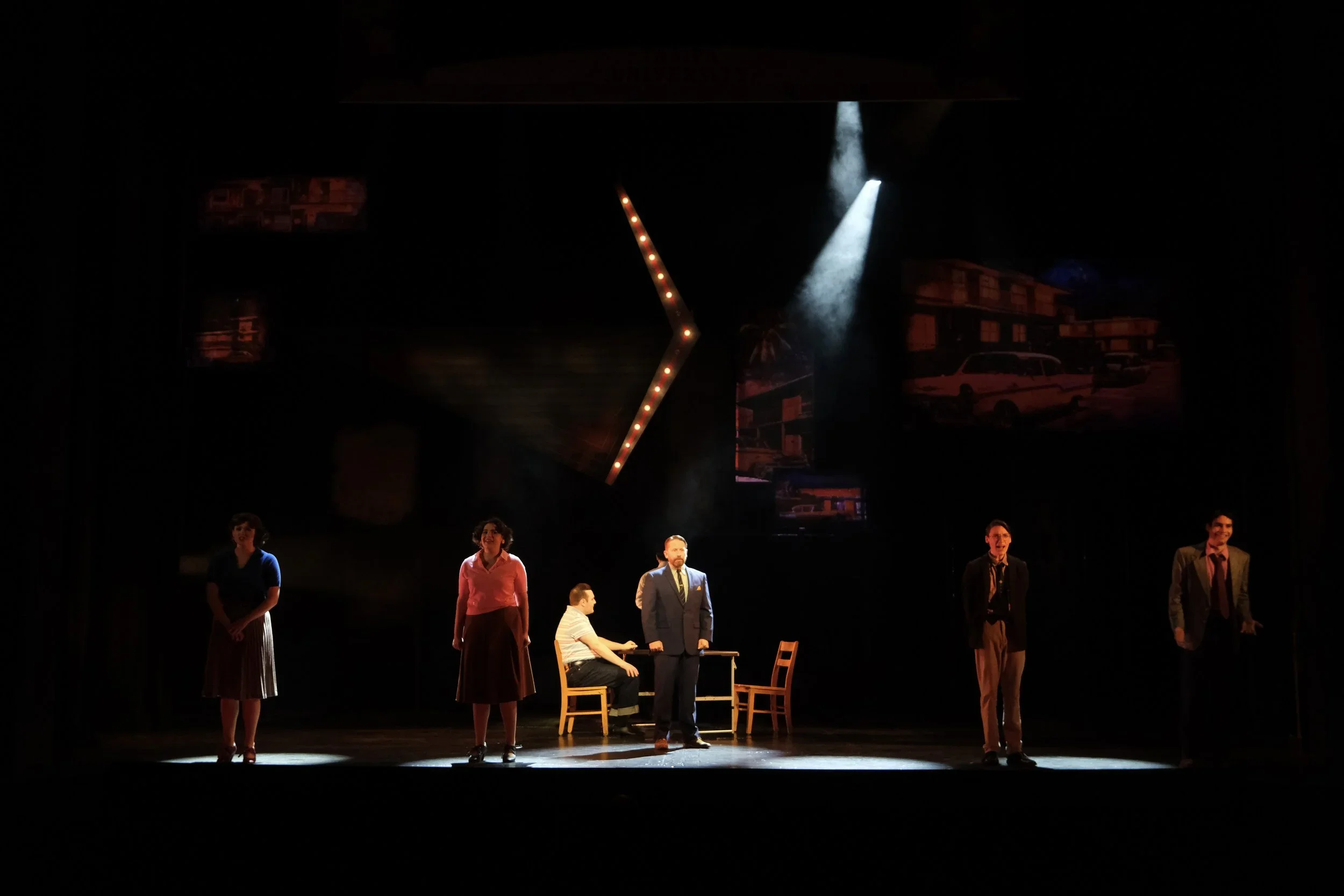 Spotlit actors facing the audience under a hanging lit up arrow set piece onstage for the world premiere musical “In a Sunshine State.”
