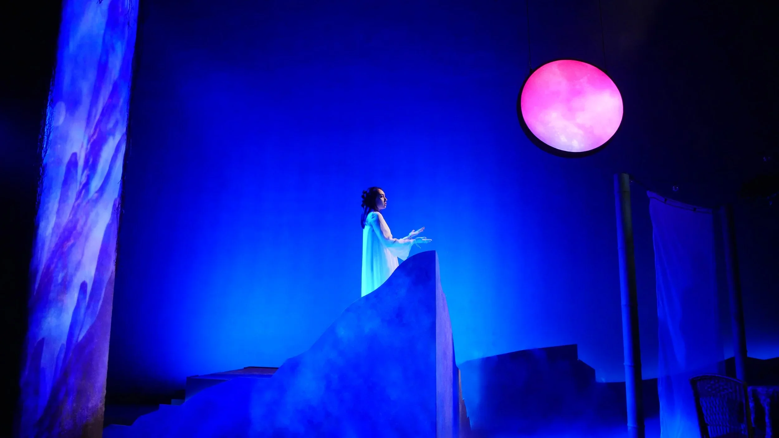Actress standing on a cliff highlighted by a glowing moon and tree and surrounded by haze on the scenic design for the play “Bloodletting” by Boni Alvarez