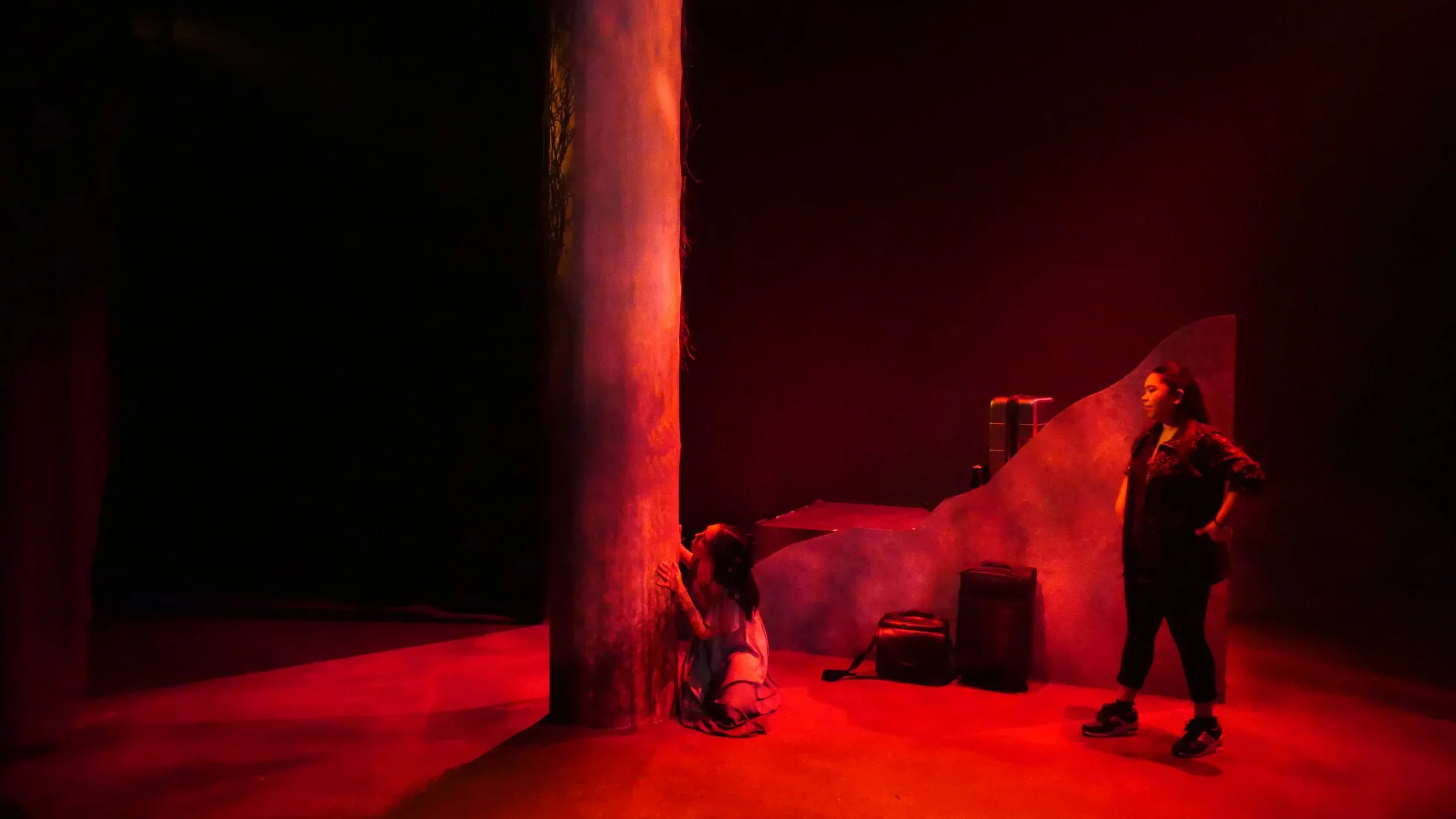 Red lit shot of Farrah and LeeLee looking at the tree on the set design of the play “Bloodletting” by Boni Alvarez