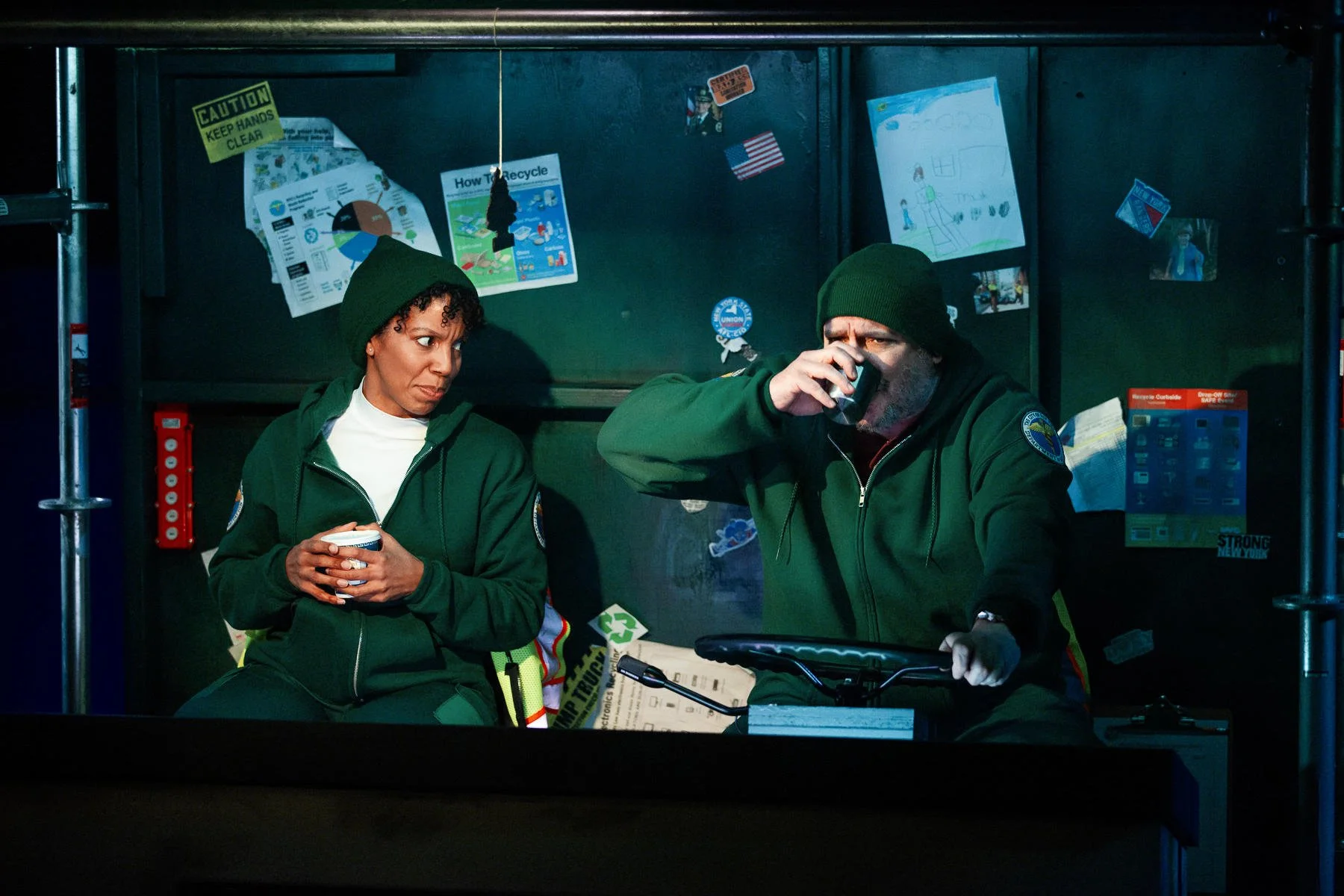 The actors in the cabin of the garbage truck drinking coffee and surrounded by graphic designed set dressing, in the play “The Garbologists” by Lindsay Joelle