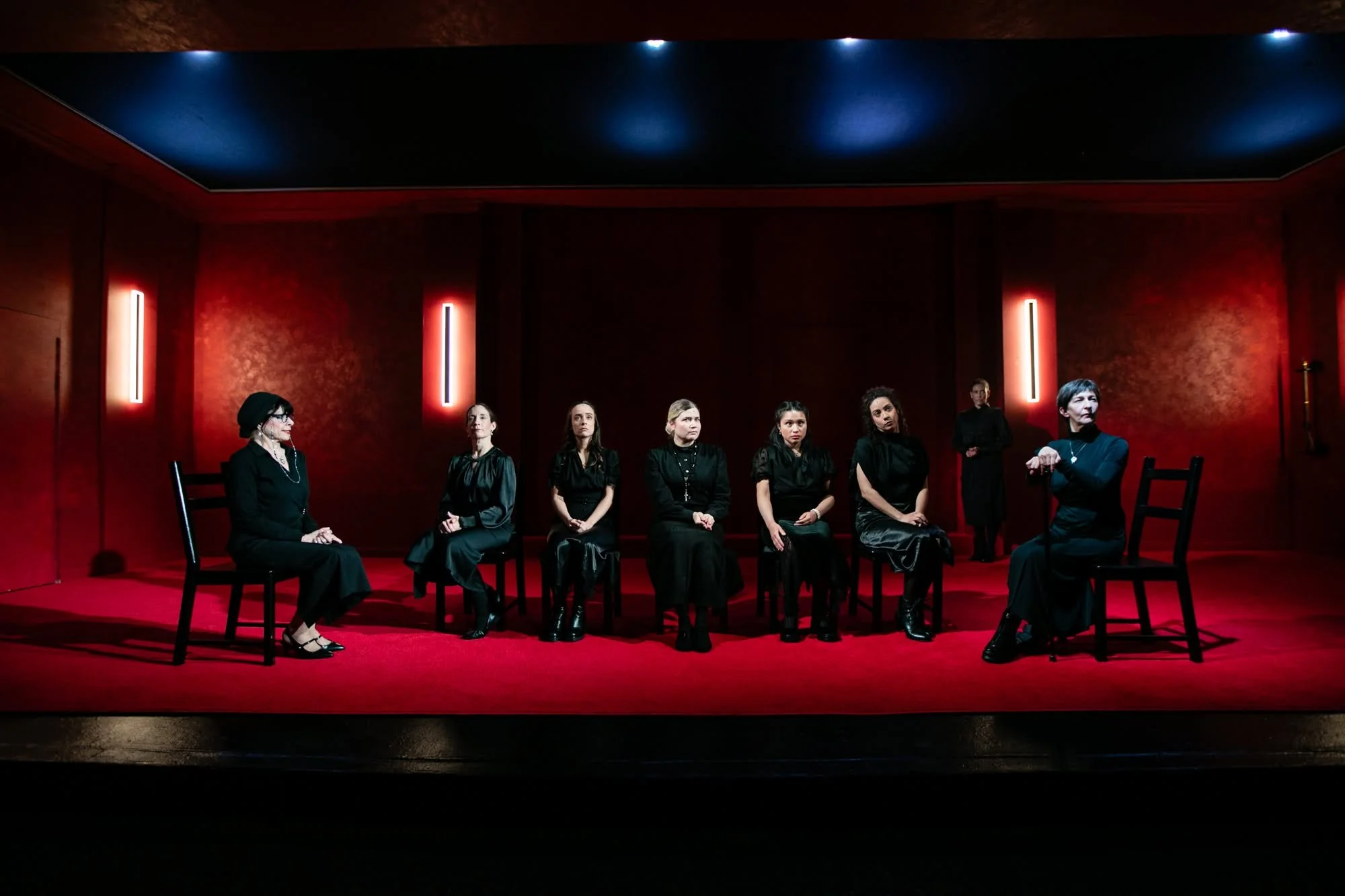 Wideshot of the all women cast seated in painted black chairs for the play “La Casa de Bernarda Alba” by Federico García Lorca