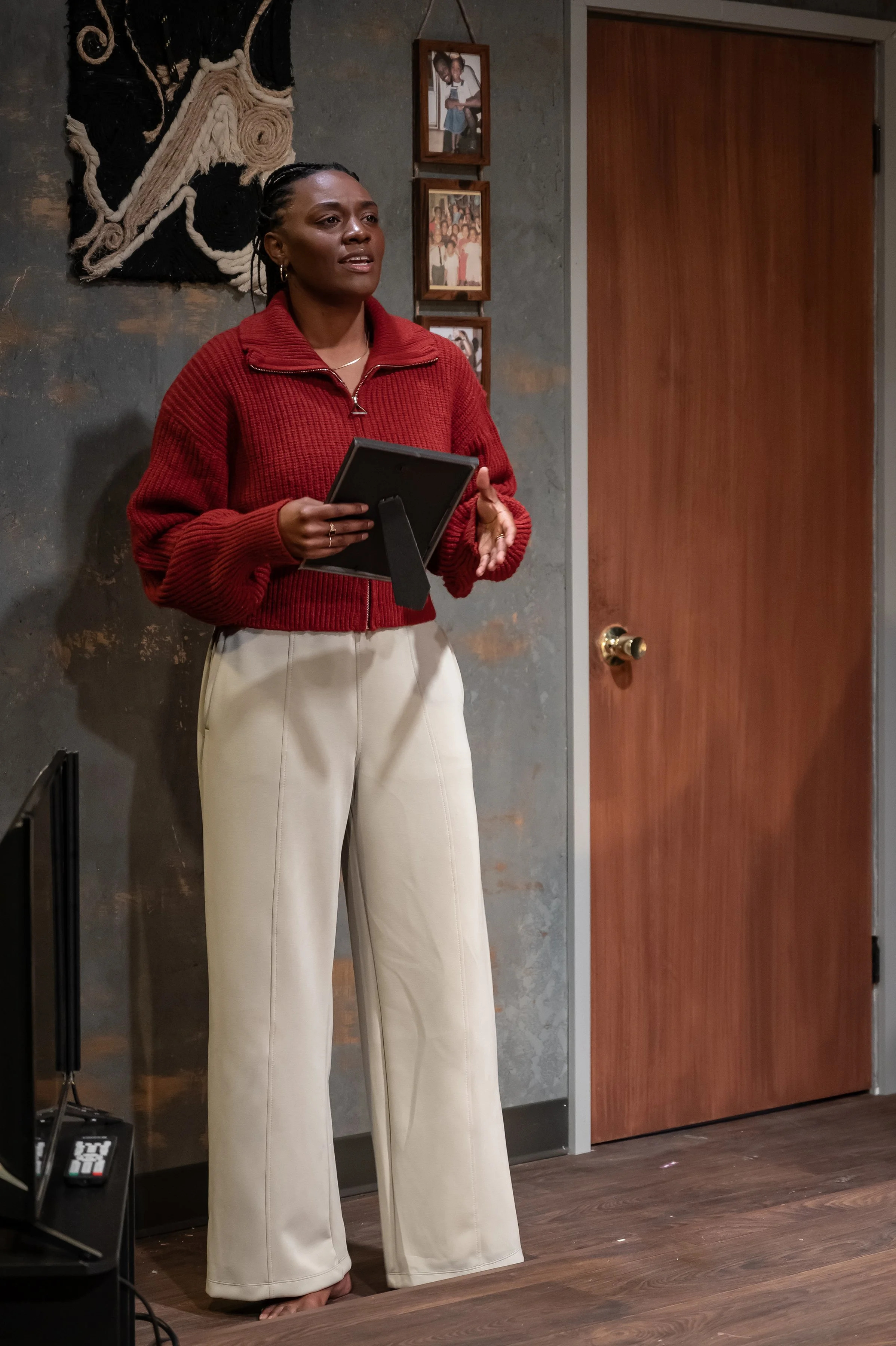 Close-up of Mina against her apartment wall dressed with a handmade rope art piece and family photos in the play “Furlough’s Paradise” by A.K. Payne