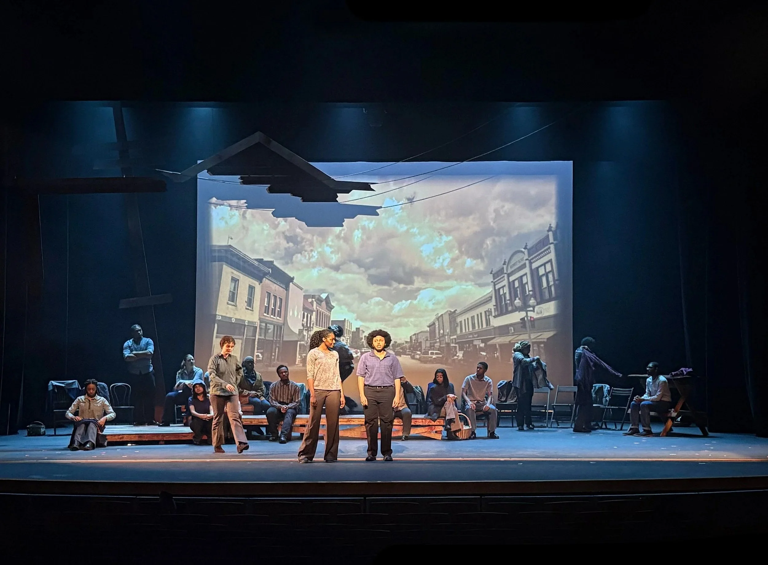 Actors facing the audience in front of the town projection in the play “The Laramie Project” by Moises Kaufman and the Tectonic Theater Project