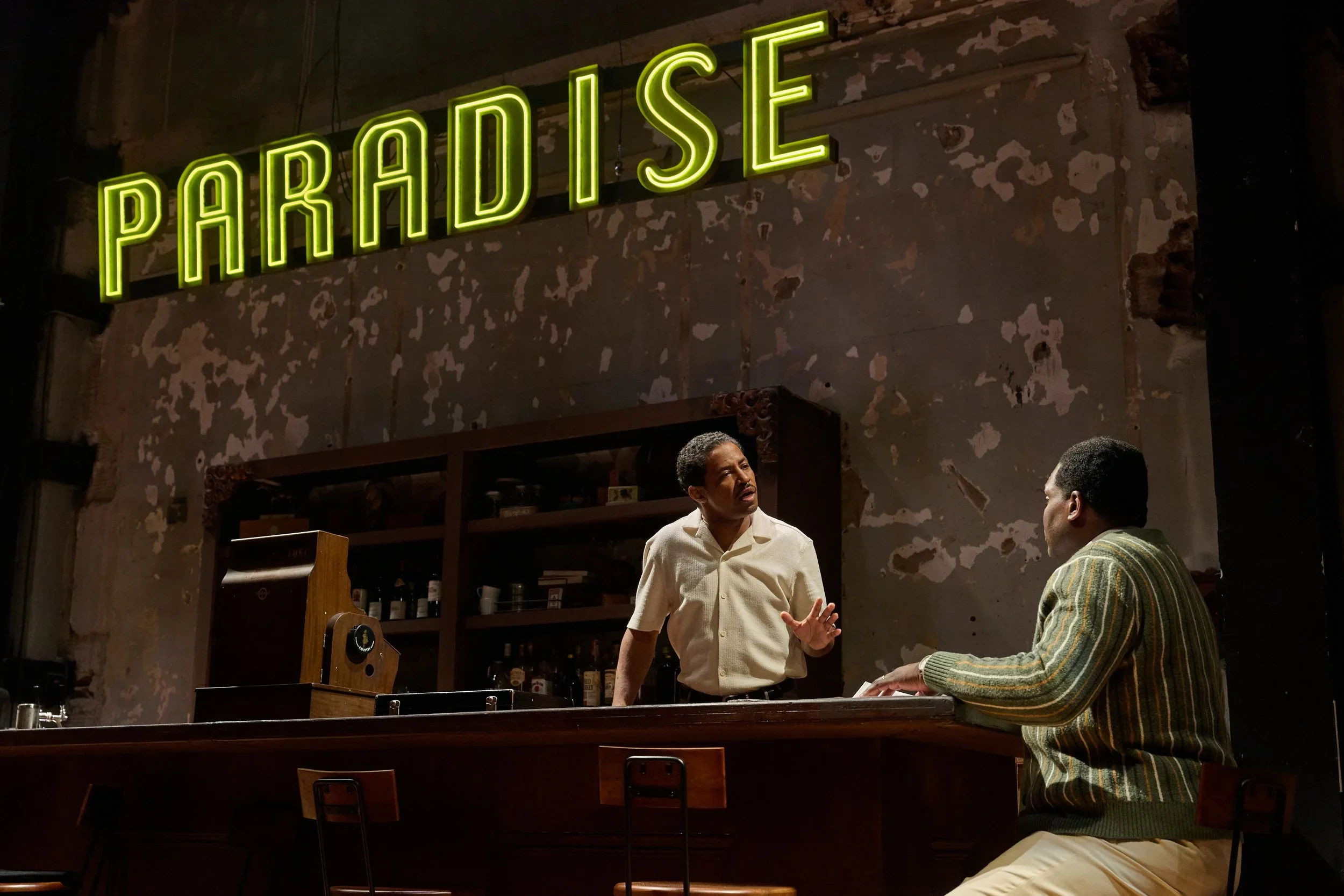 Two men are having a conversation in a dimly lit bar or restaurant with a neon green sign that says 'PARADISE' on the wall behind them.