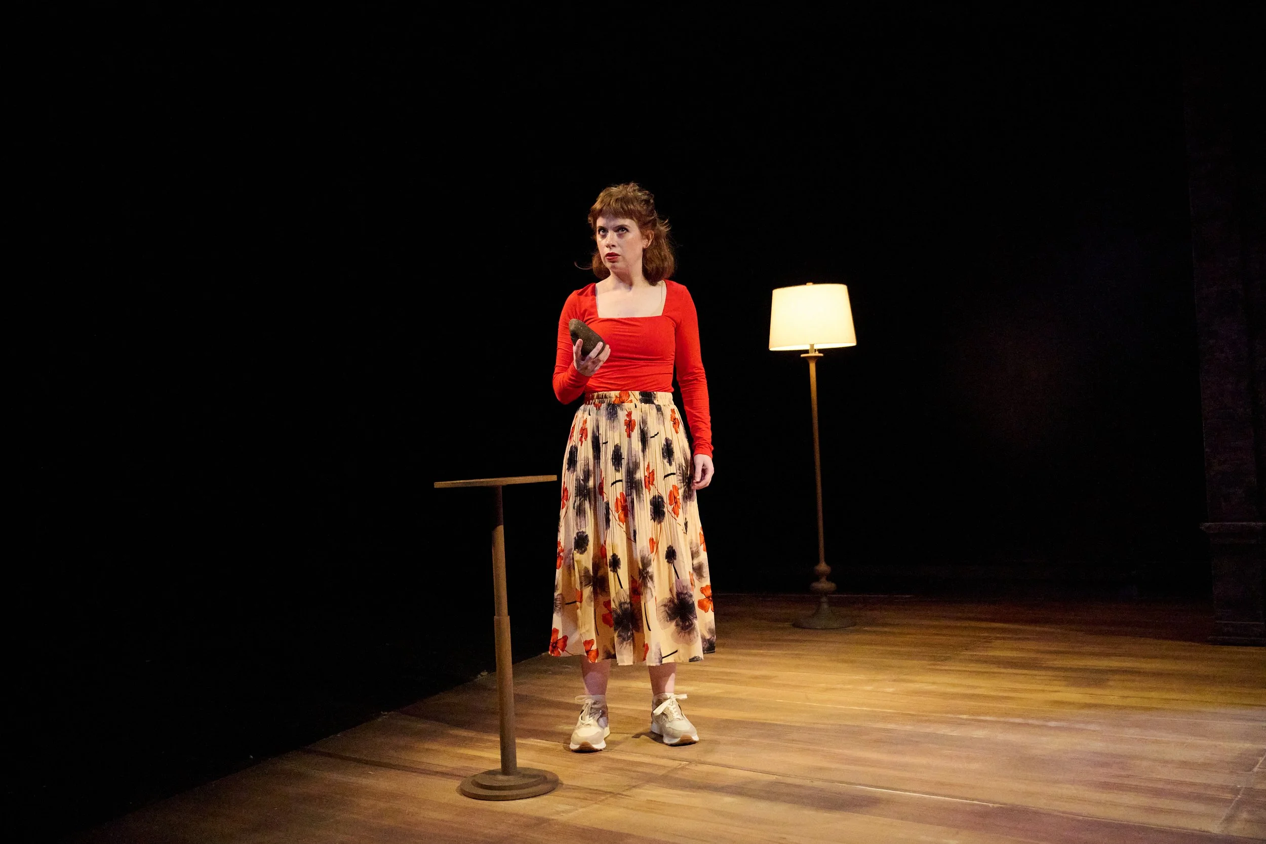 A woman onstage holding a rock by a small table and floor lamp in the North American premiere of “Lie Low” by Ciara Elizabeth Smyth