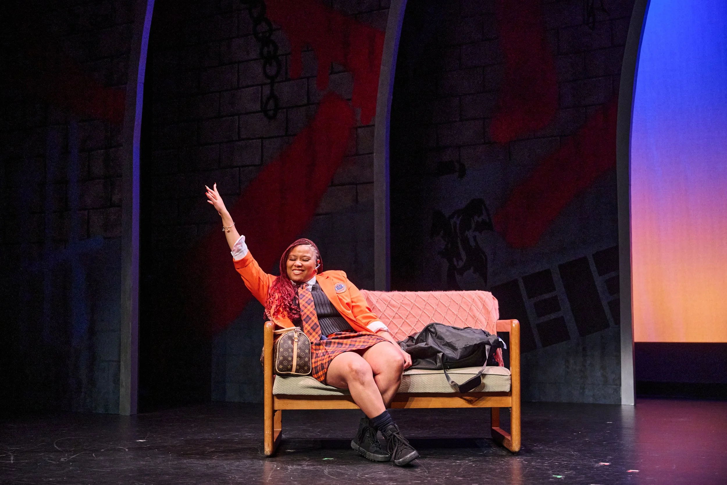 A woman with braided hair wearing an orange blazer, plaid dress, and black shoes, sitting on a chair on stage with a background featuring dark banners with red and black designs. She is smiling, with her left arm raised, and has a small handbag and a bag on the floor next to her.