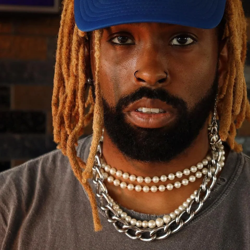 Close-up of a man with blond dreadlocks wearing a blue cap, silver hoop earring, multiple pearl necklaces, and a chunky silver chain, standing in front of a brick wall. Myc Daz.