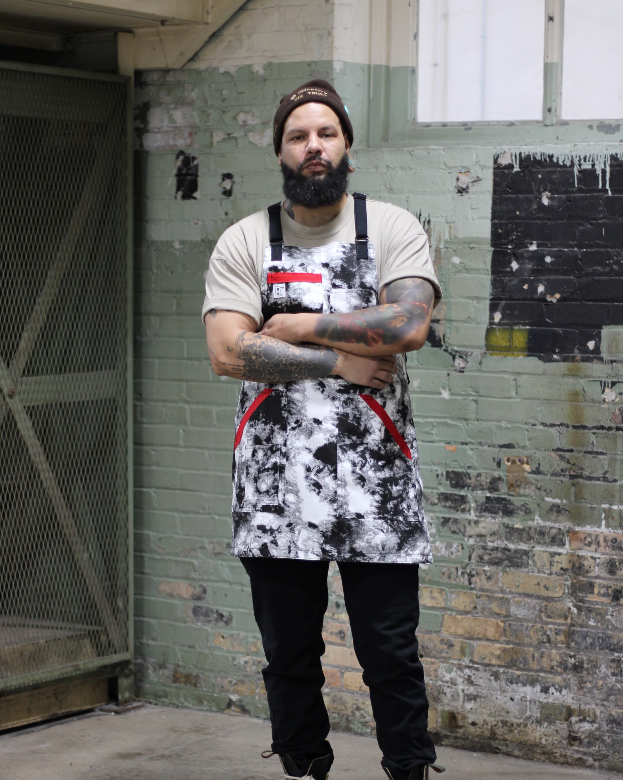 A man with a beard, tattoos, and wearing a beanie, T-shirt, and apron standing with arms crossed in front of a painted brick wall with peeling paint and an open window.