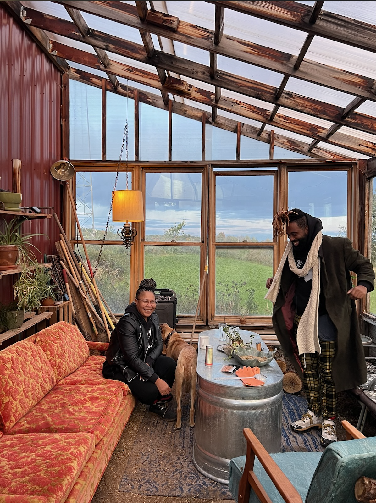 Two people gather inside a rustic greenhouse with large windows. One woman sitting on a red patterned couch is smiling and petting a dog, while a man standing nearby is smiling and adjusting his coat. The space has plants, gardening tools, and a warm lamp hanging from the ceiling, with a view of green fields outdoors.