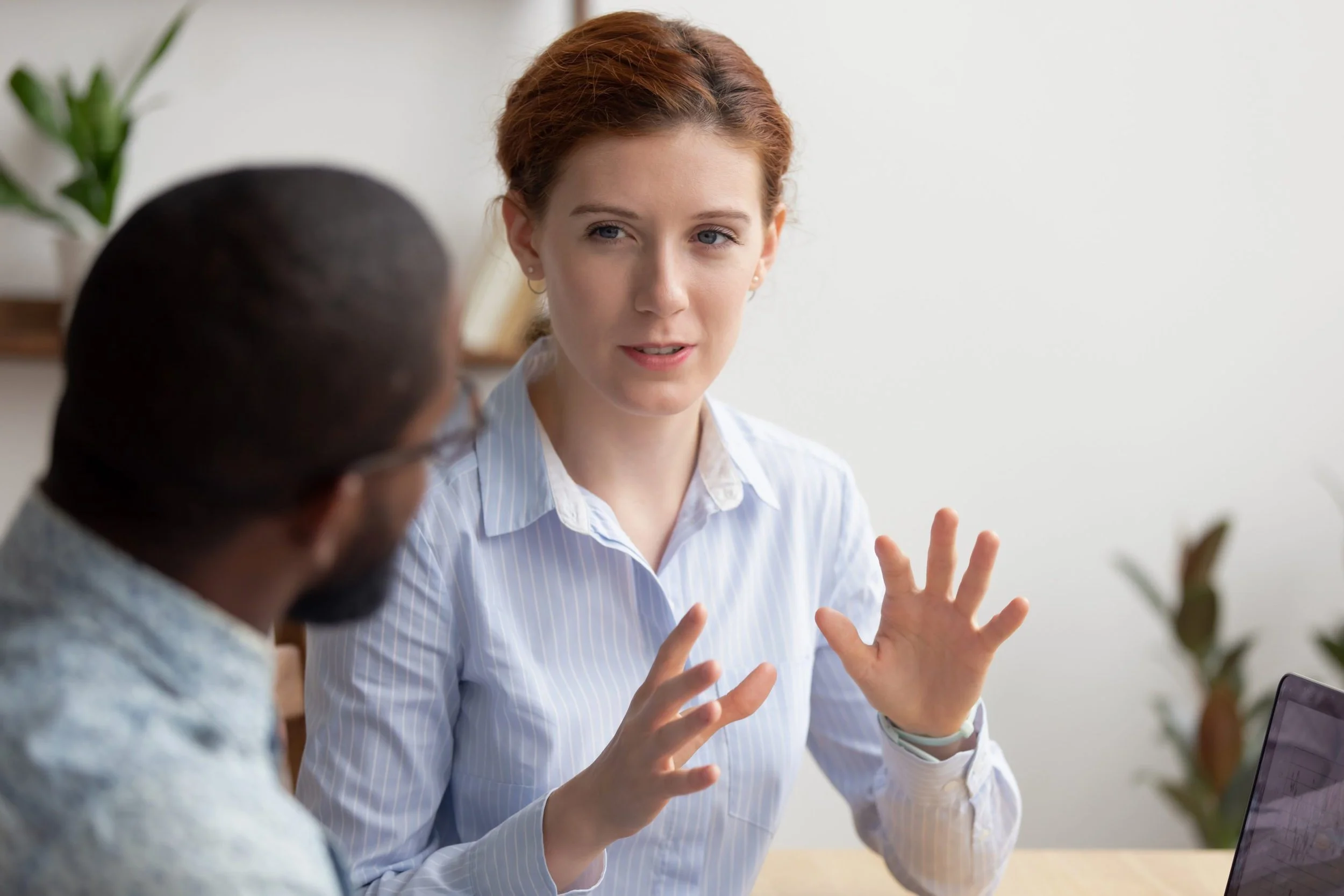 A woman with red hair and blue eyes is engaged in a conversation with a man. She is gesturing with her hands while speaking, and they are seated at a desk with a laptop visible. The background is a plain white wall with a plant on the left.
