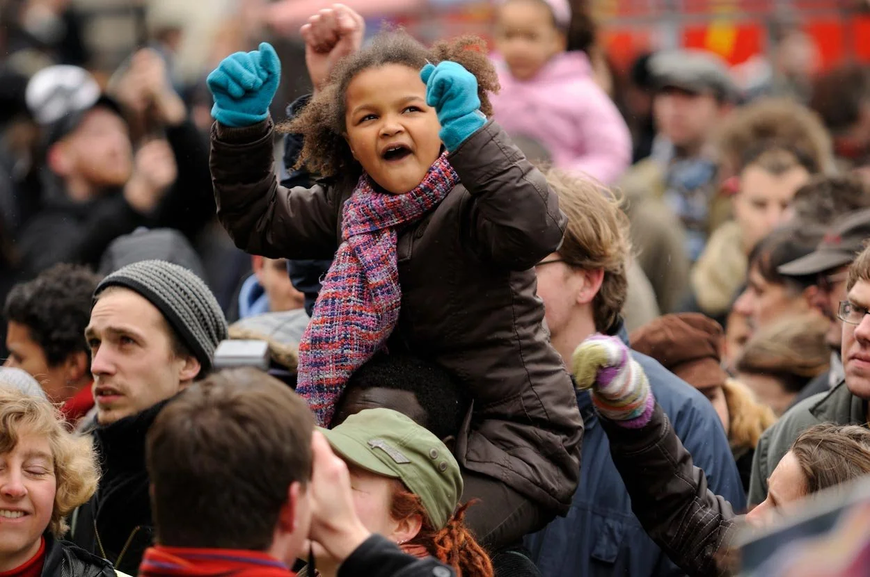 Young girl with curly hair wearing a scarf and blue gloves sits on someone's shoulders during a crowded outdoor event, expressing excitement.