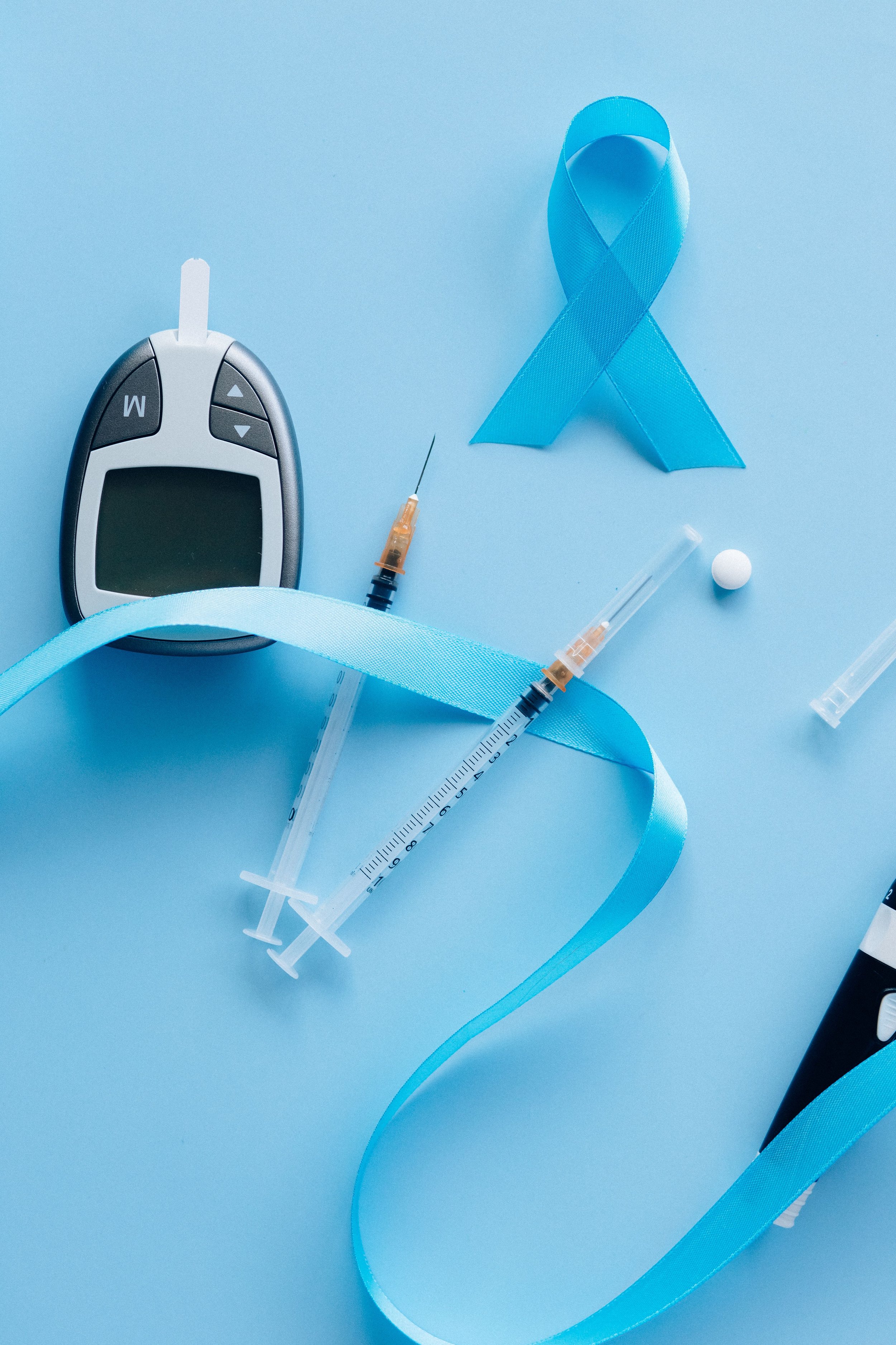 A blue awareness ribbon, a blood glucose meter, a syringe, a pill, and medical supplies placed on a blue background, representing diabetes awareness.