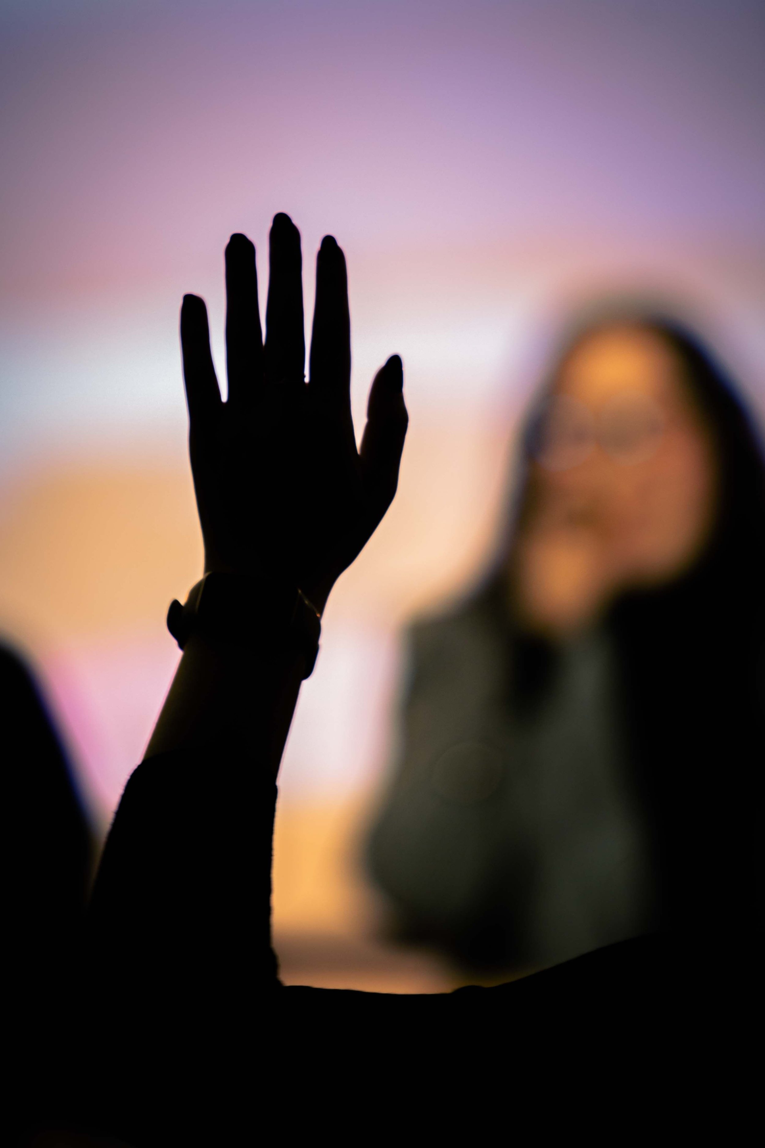 Silhouette of a hand with open fingers raised against a blurred sunset or sunrise sky.