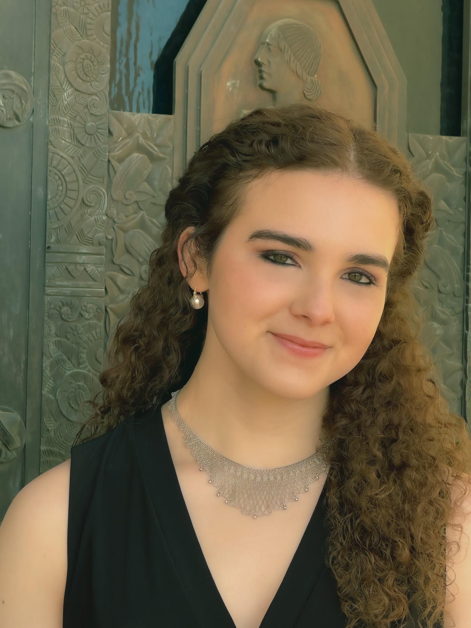 Close-up of a young woman with curly hair and green eyes, wearing pearl earrings, a diamond necklace, and a black sleeveless dress, standing in front of an ornate metal door with relief designs and a profile face bas-relief.