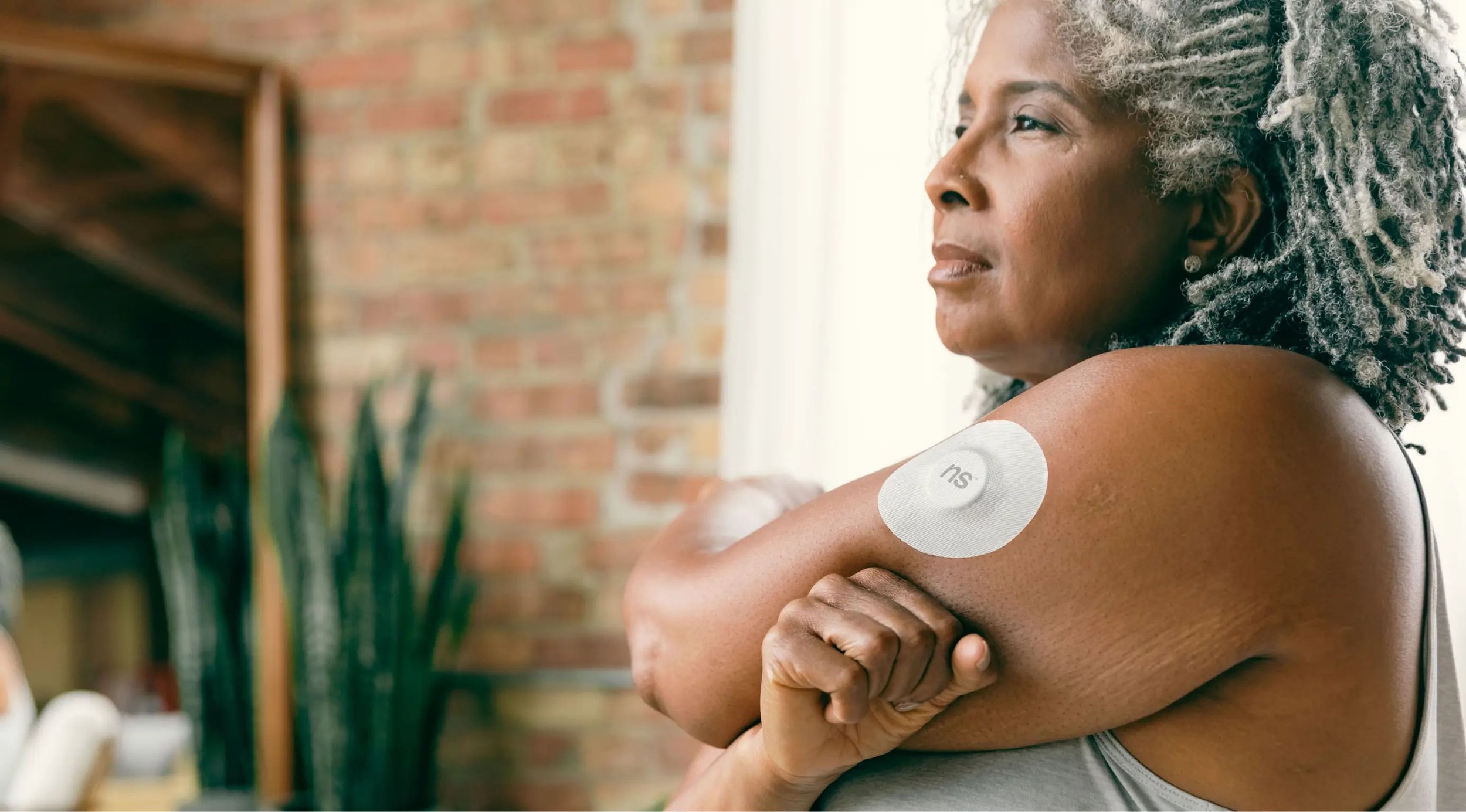 A woman with curly gray hair applying an adhesive patch labeled 'SU' to her upper arm, looking content while sitting indoors with a brick wall and plants in the background.