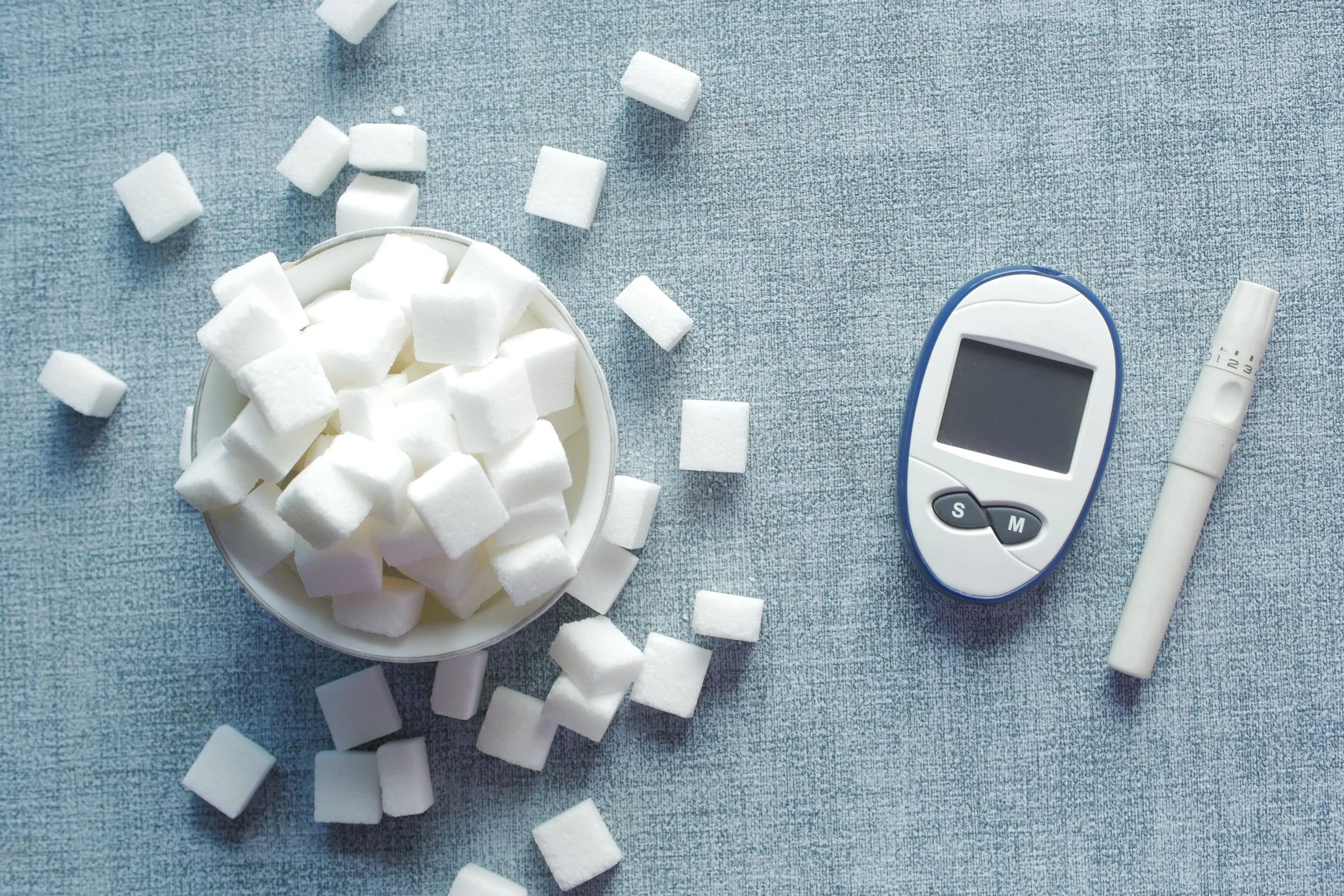 A bowl of sugar cubes on a blue surface next to a blood glucose meter, a white pen-like device, and a white marker.